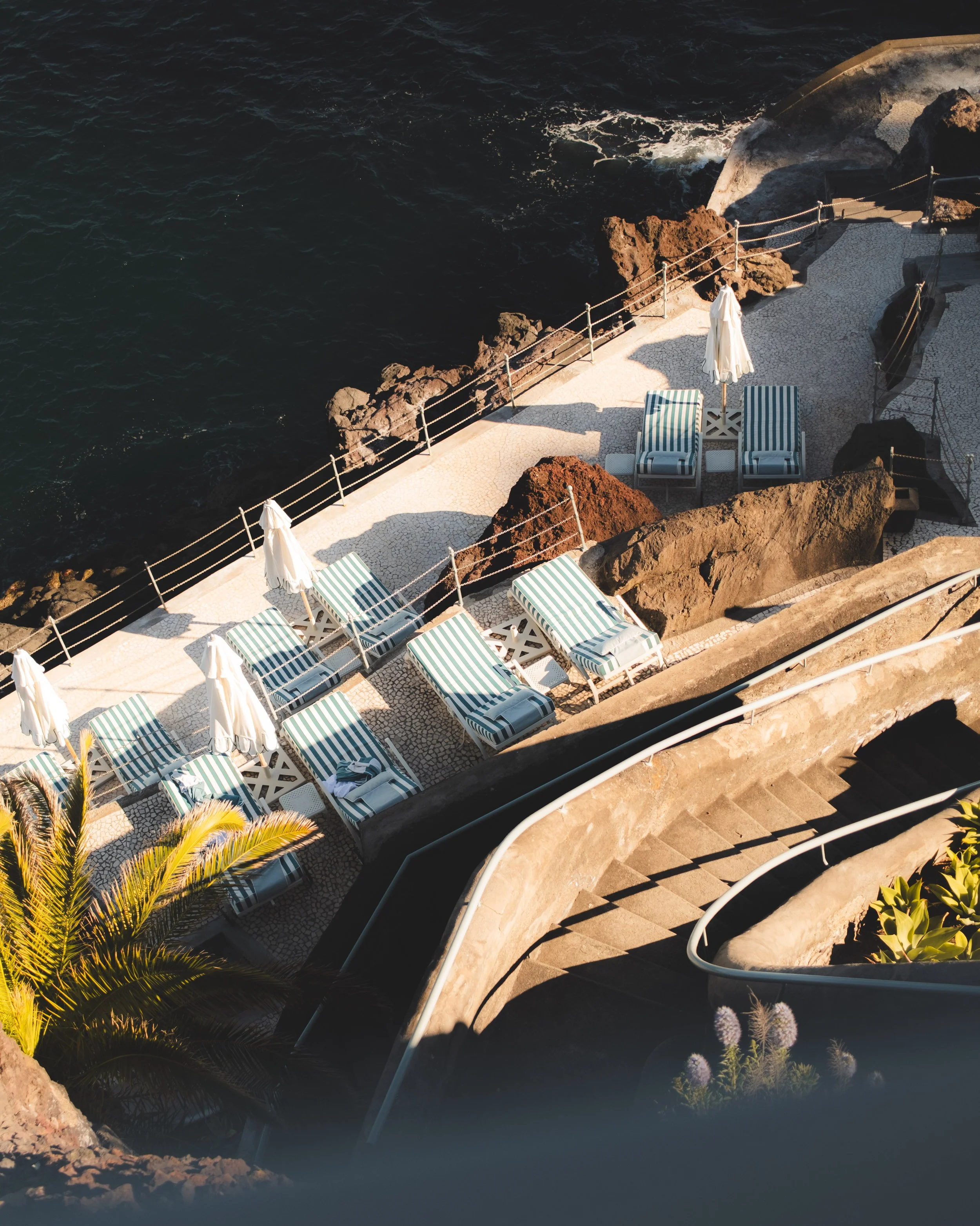 Seaside area with turquoise striped lounge chairs and white umbrellas on a shaded stone patio overlooking the ocean, with rocks and coastal plants.