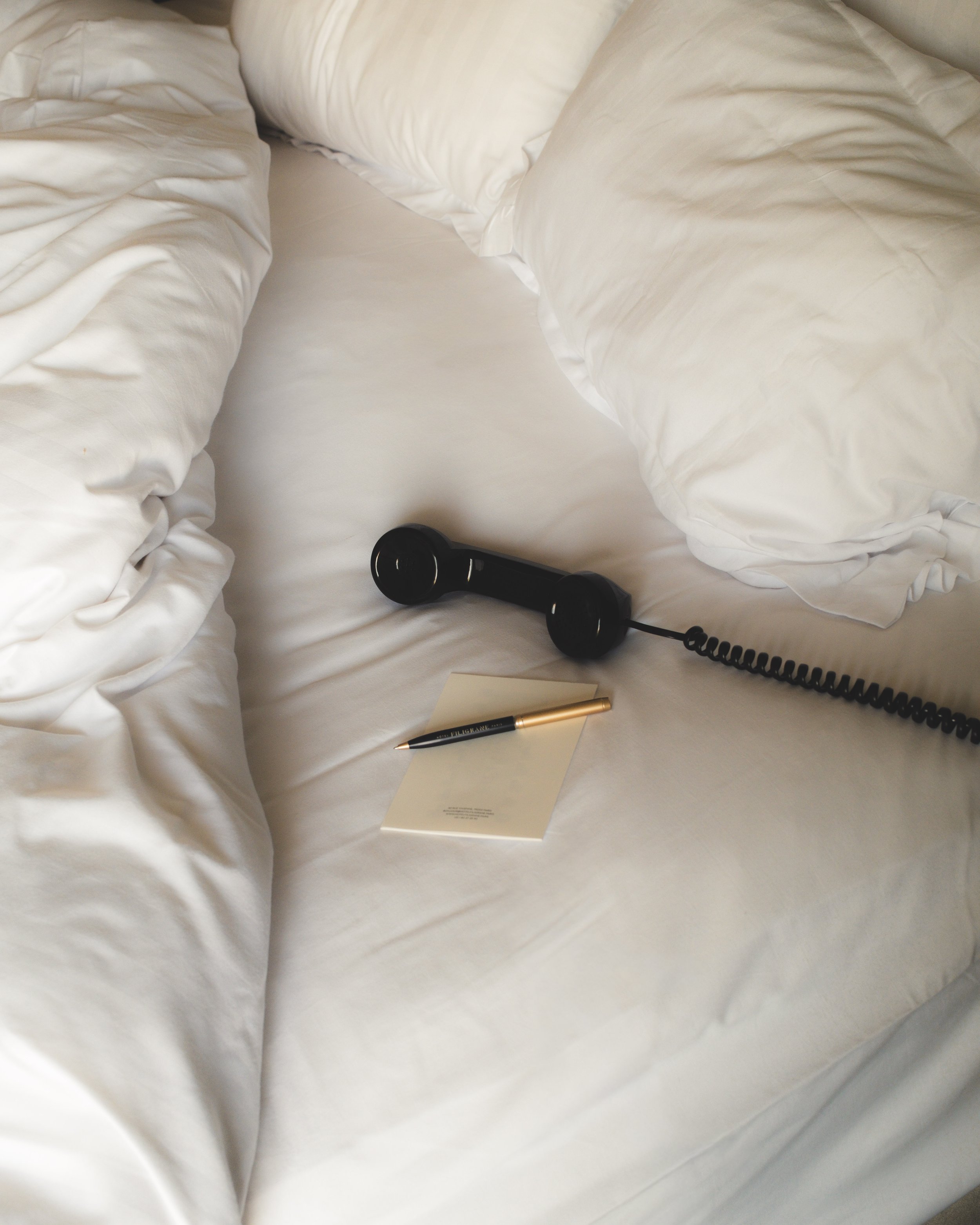 Close-up of a white bed with two white pillows, a black phone, a gold pen, and a small notepad resting on the bed sheets.