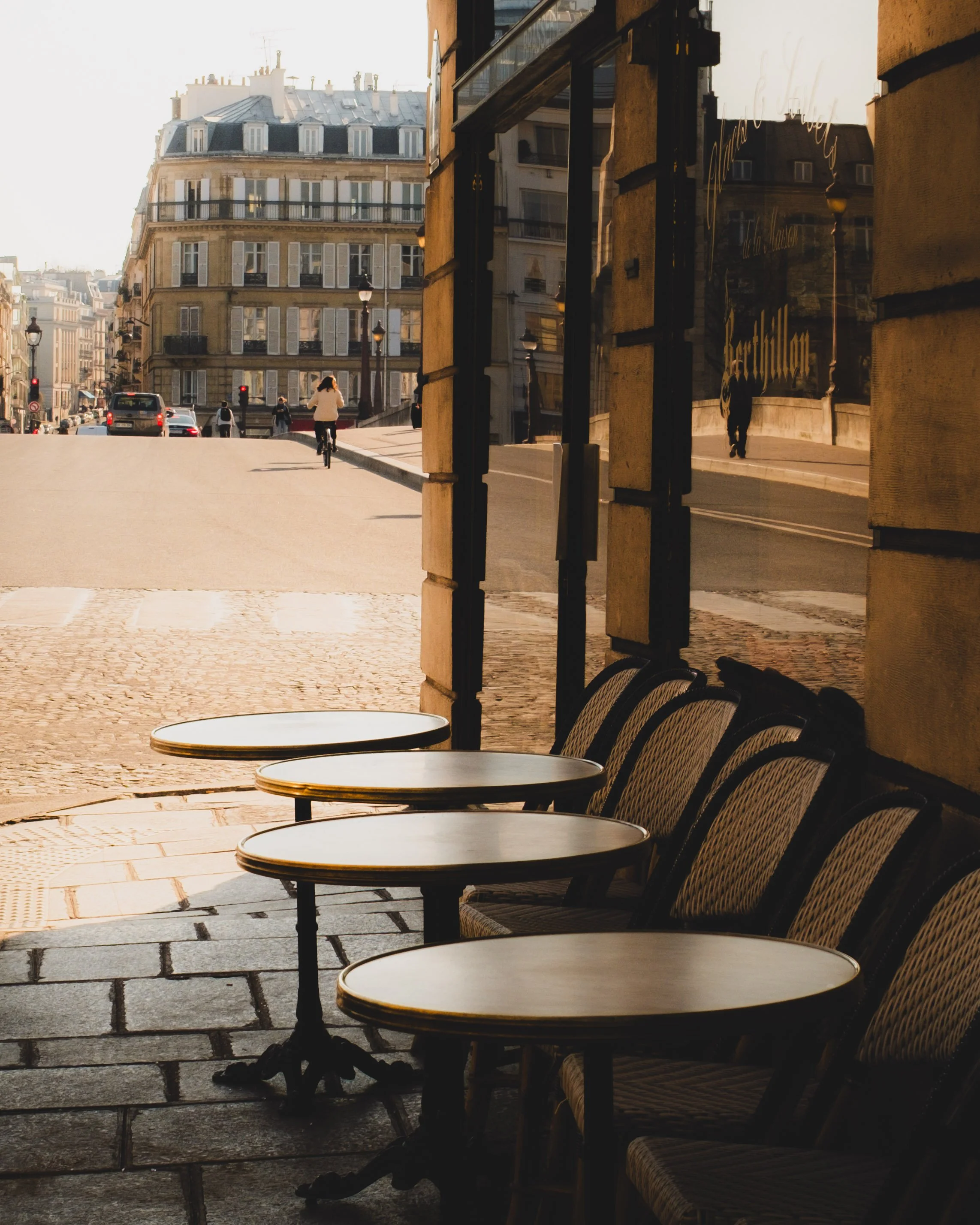 Outdoor cafe tables and chairs on a cobblestone street in Paris, with classic Paris buildings in the background and a cyclist riding by. Lionel Chu - Hotel & Travel Photographer.