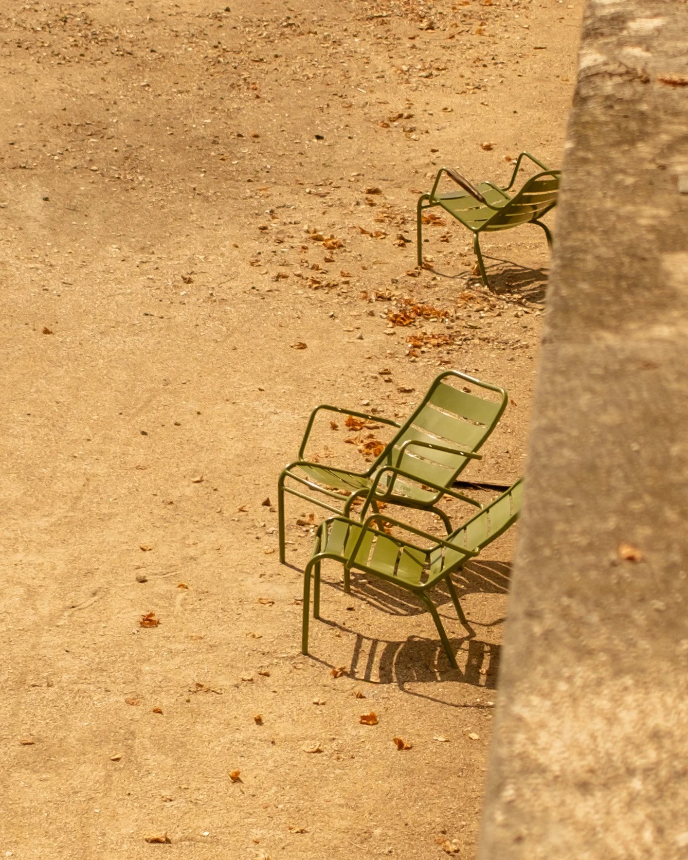 Three empty green chairs on brown sandy ground with long shadows and a blurred stone foreground. Jardin des Tuileries, Paris. Lionel Chu - Hotel & Travel Photographer.
