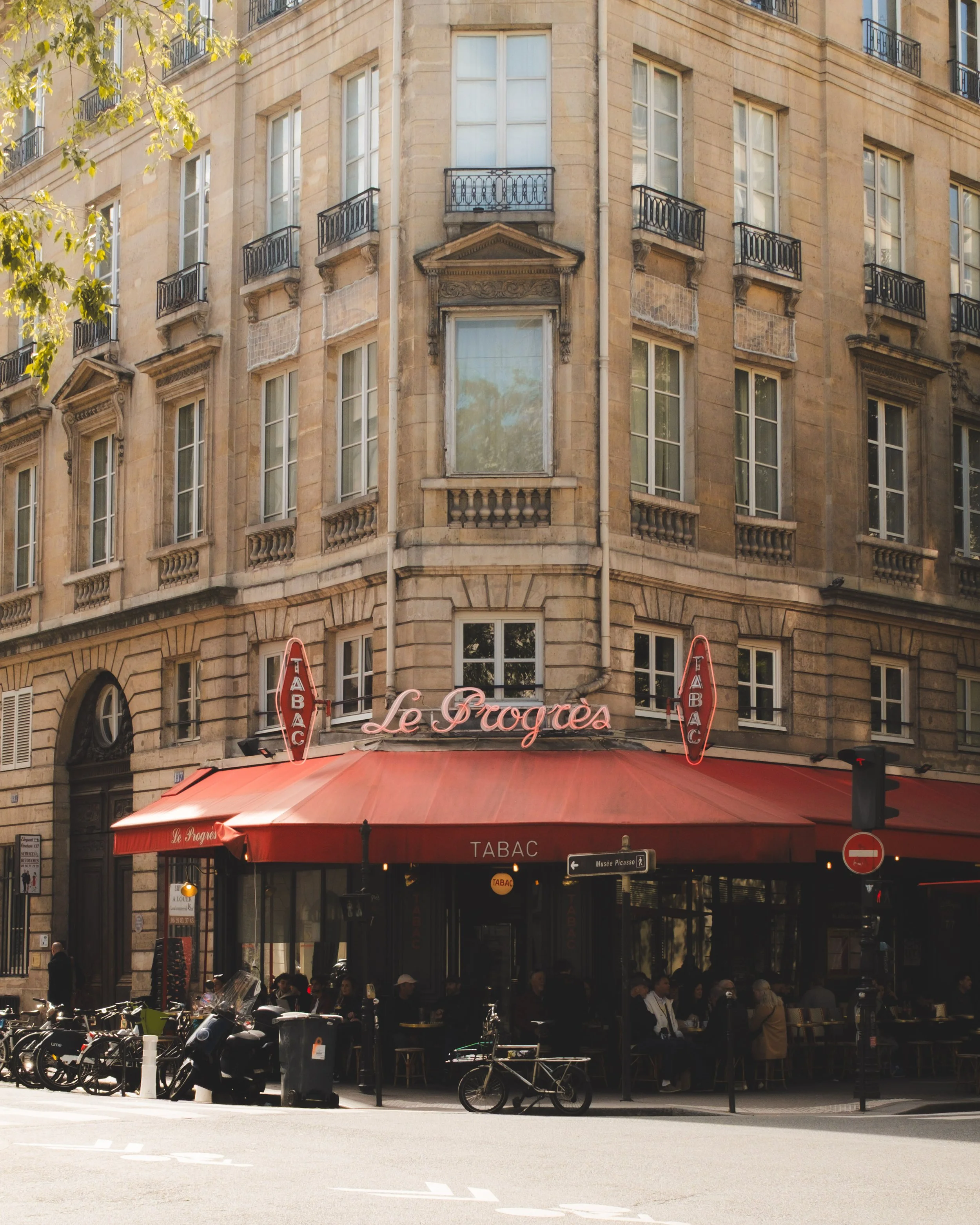 A beige stone building with multiple windows and small black balconies. The ground level features a restaurant or cafe named Le Progrès with a red awning and outdoor seating. Several bicycles are parked in front, and people are sitting at tables or w
