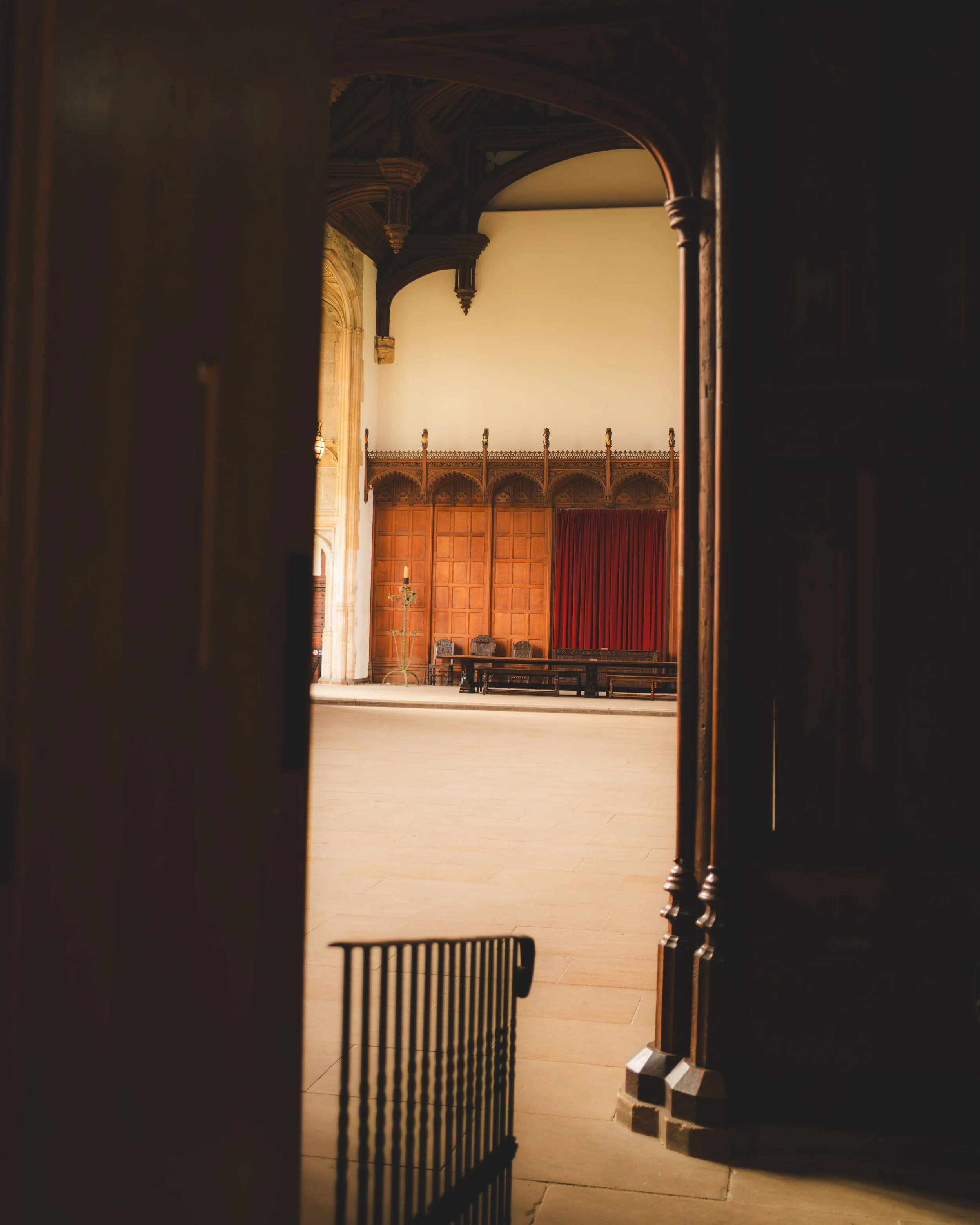 The image shows the interior of a large, historic room or hall, viewed through an open doorway. The room features wooden paneling on the walls, an ornate wooden balcony or gallery with decorative carvings, a red curtain, and rows of chairs. The ceili