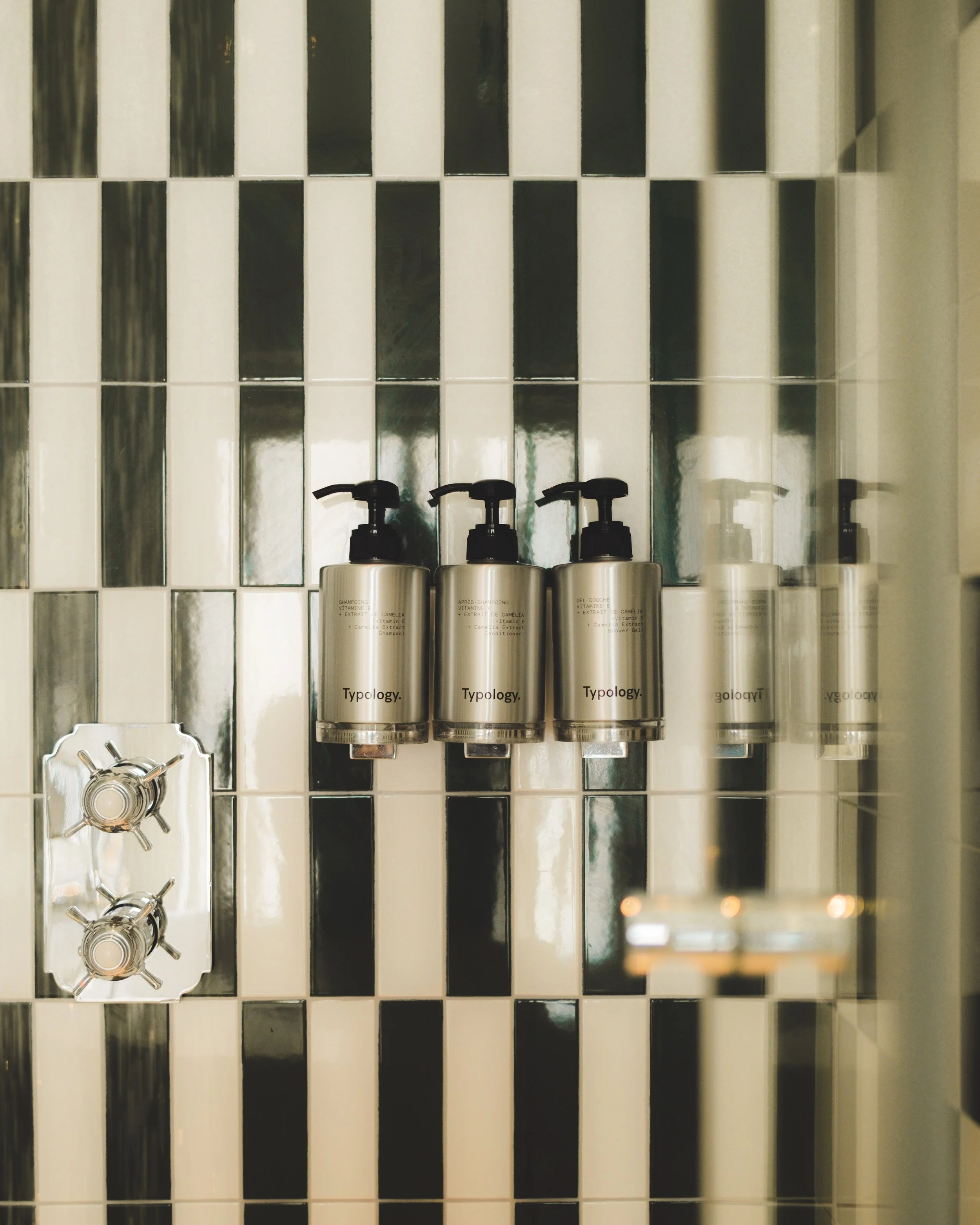 Four amenities bottles with black pump dispensers on a black and white tiled bathroom wall, with a silver faucet below.