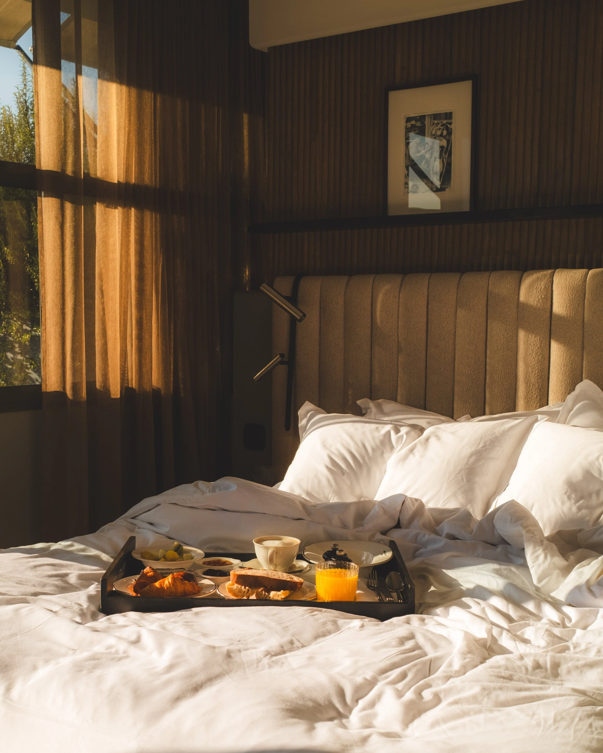 Breakfast tray with croissant, bread, butter, juice, and other items on a white bed with sunlight coming through a window and brown curtains, in a bedroom with wood-paneled wall and framed artwork.
