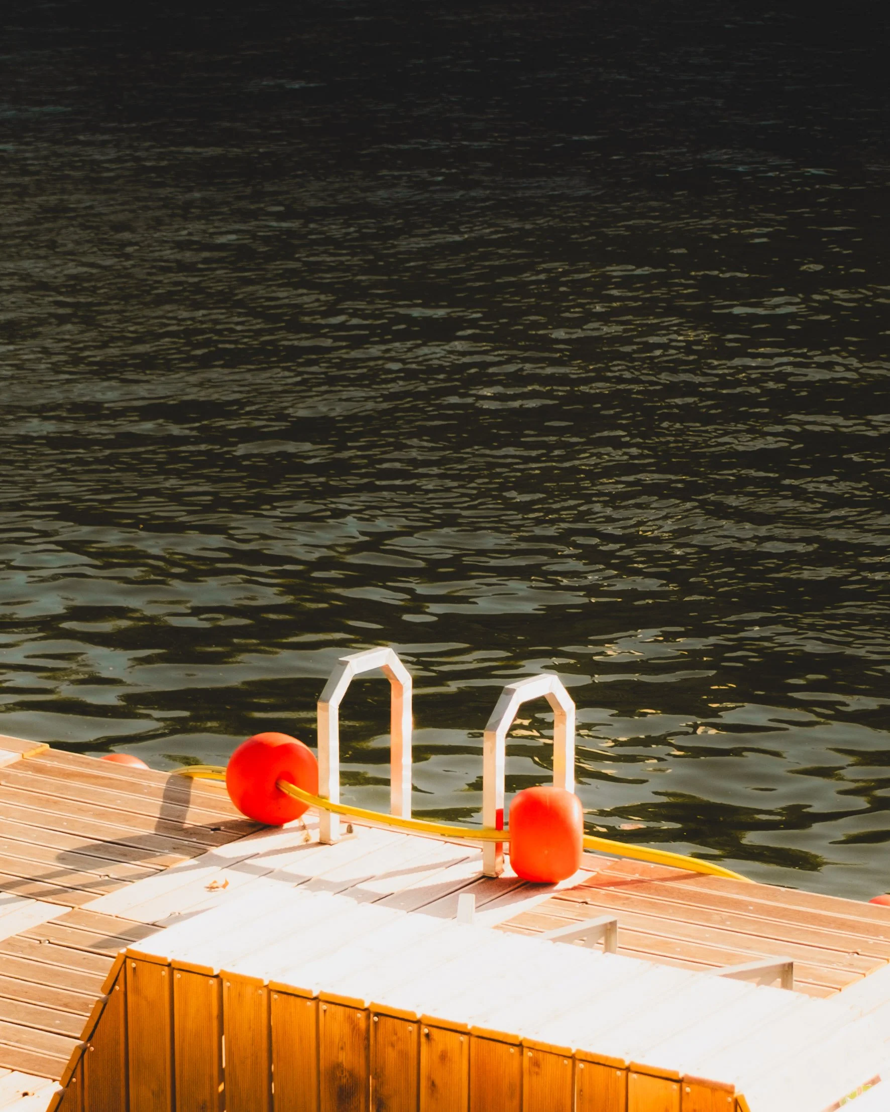 A wooden dock with metal handrails, orange buoys, and a yellow rope against dark water at sunset. Lionel Chu - Hotel & Travel Photographer