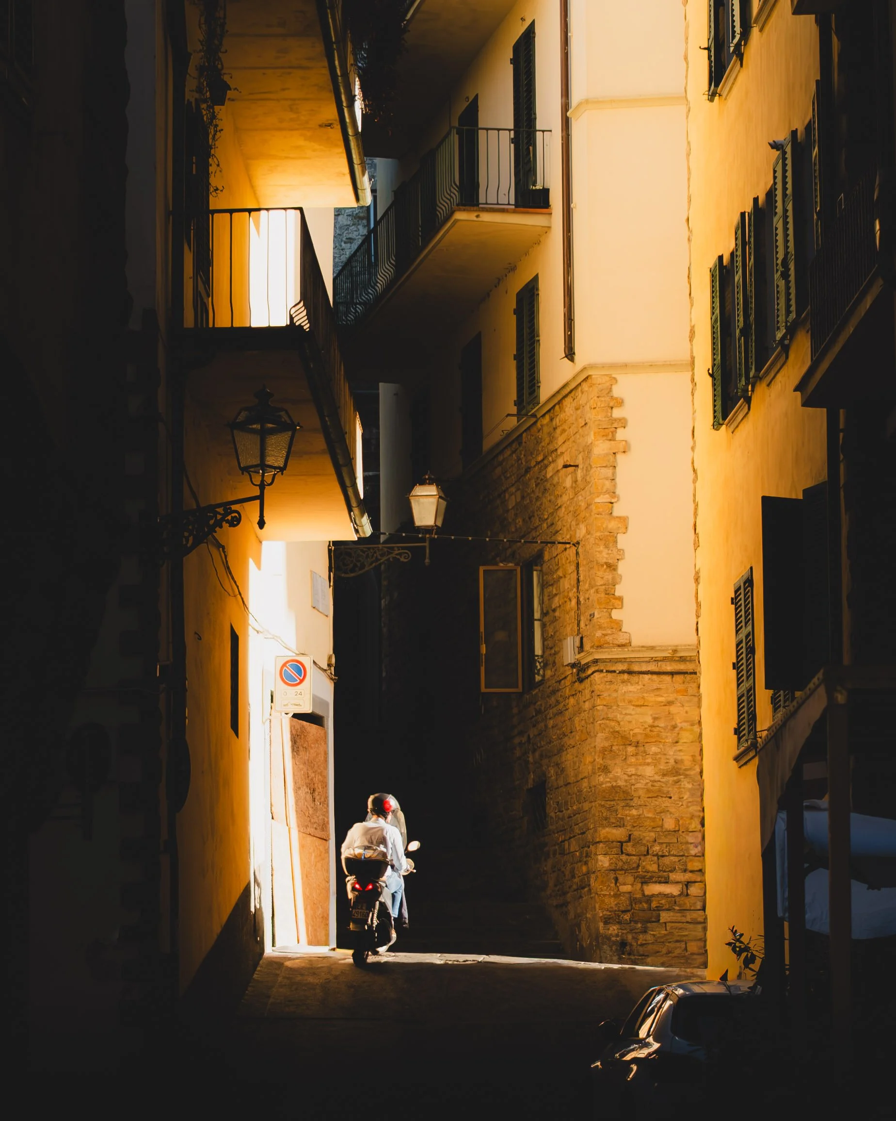 A narrow street in an Italian town with yellow and beige buildings, a person riding a scooter up a steep incline, and parked cars on the side. Lionel Chu - Hotel & Travel Photographer