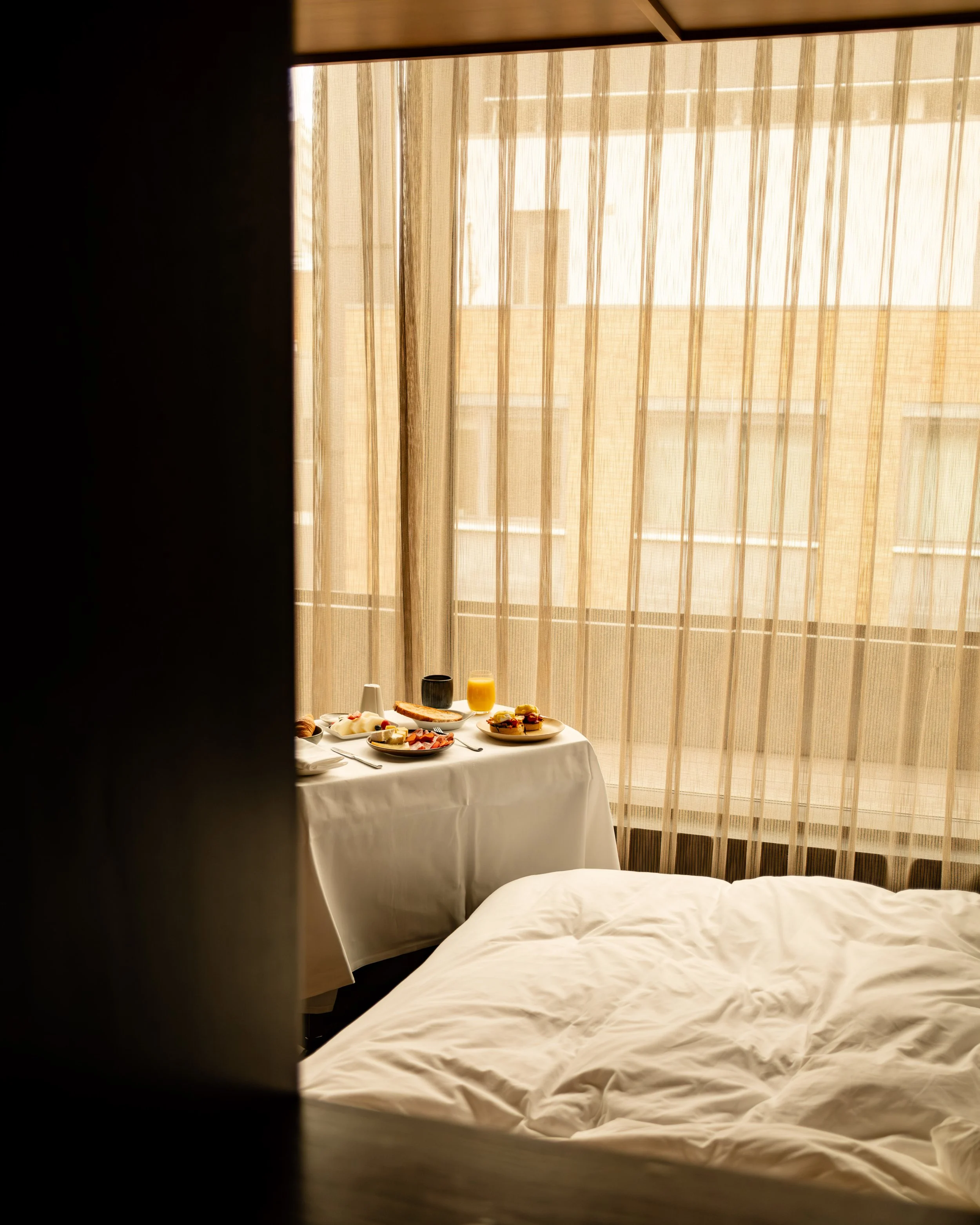 A hotel room with a bed in the foreground, a tray with breakfast on a table beside the window, sheer curtains, and a view of another building outside. Lionel Chu - Hotel & Travel Photographer