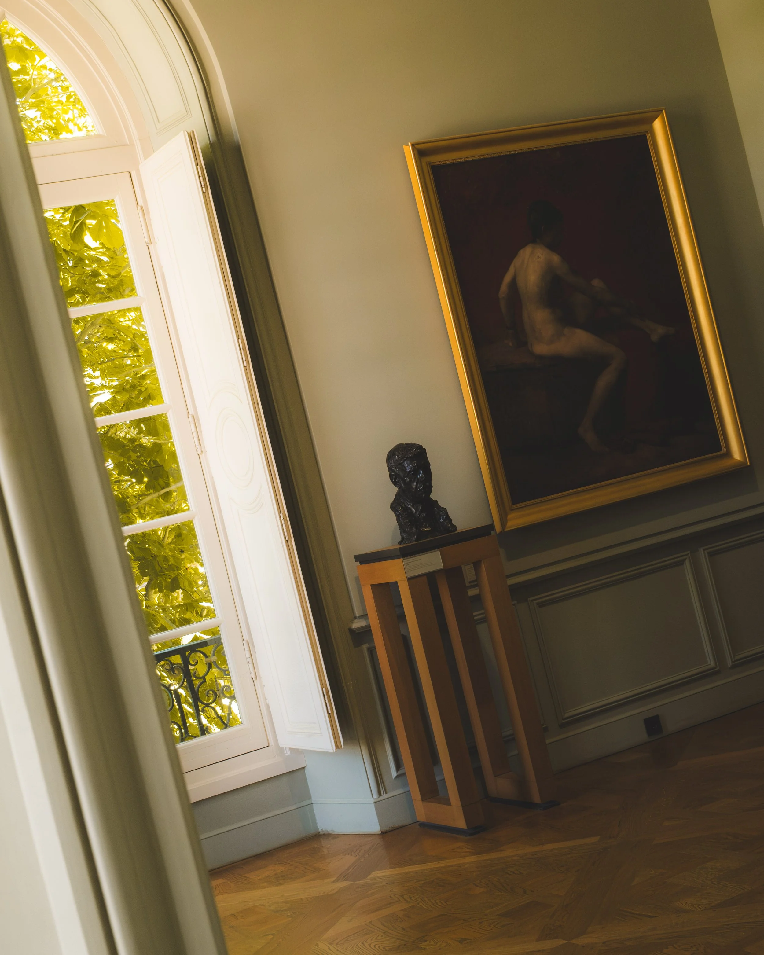 Interior of a room with a window that has white wooden shutters and yellow-green foliage outside. Inside, there is a dark framed painting of a nude figure sitting on a ledge, a sculpture bust on a stand, and small tables along the wall, with wooden f