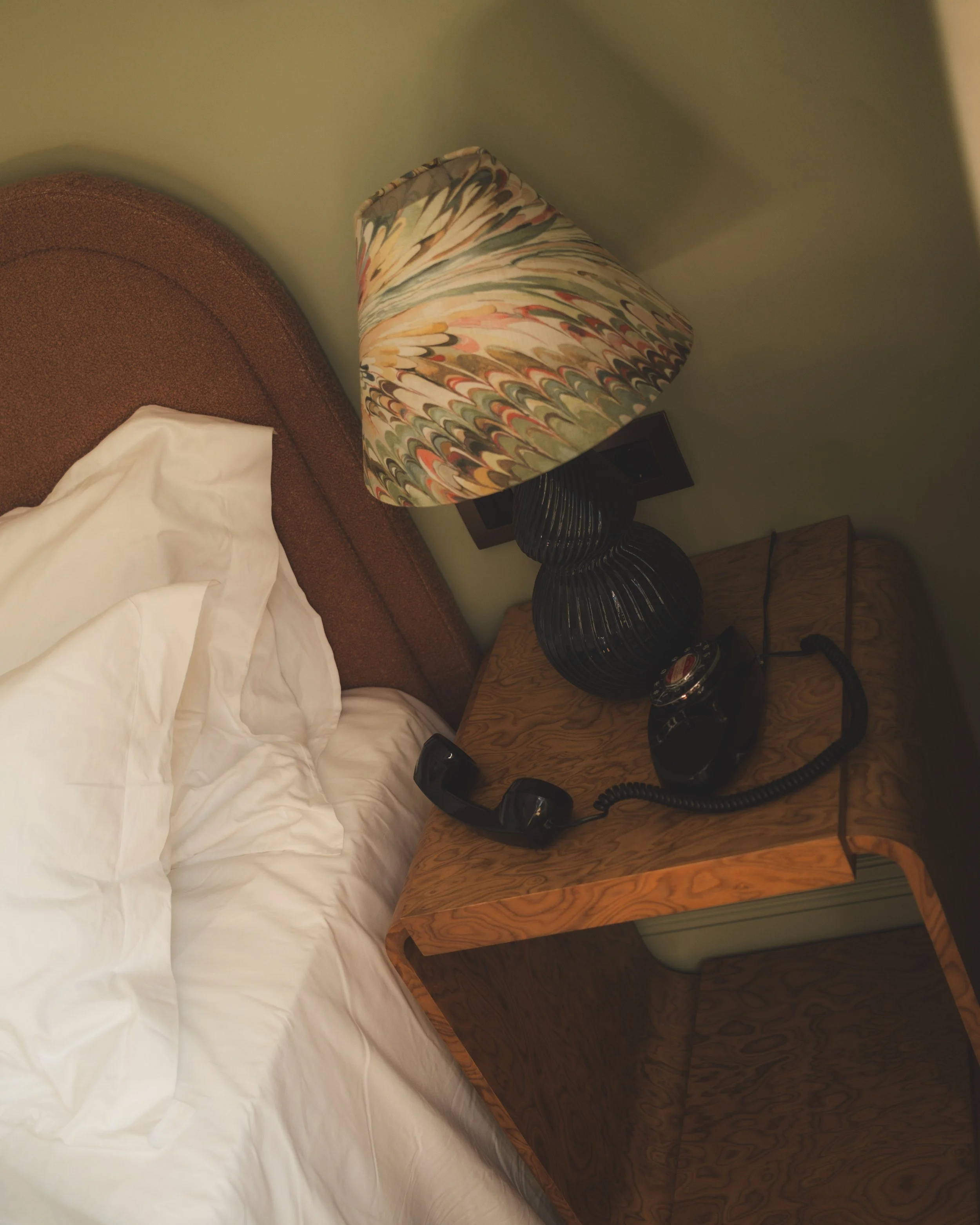 Close-up of a wooden bed and nightstand with a vintage black rotary phone, a lamp with a colorful, swirled pattern shade, and a small wooden drawer visible in a dimly lit room.