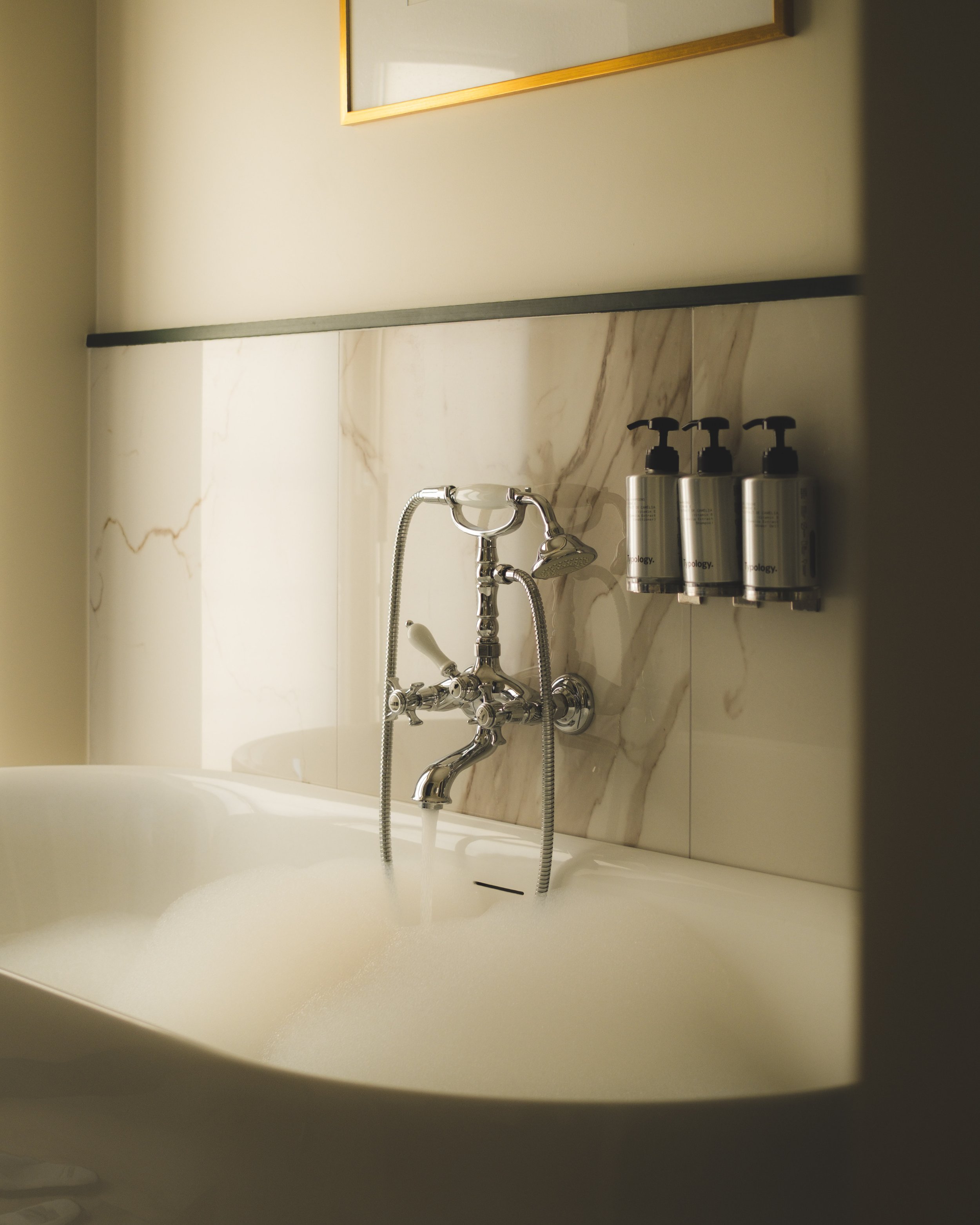Bathroom with a bathtub, gold-framed mirror, marble wall tiles, and four silver soap dispensers labeled 'Typology.'