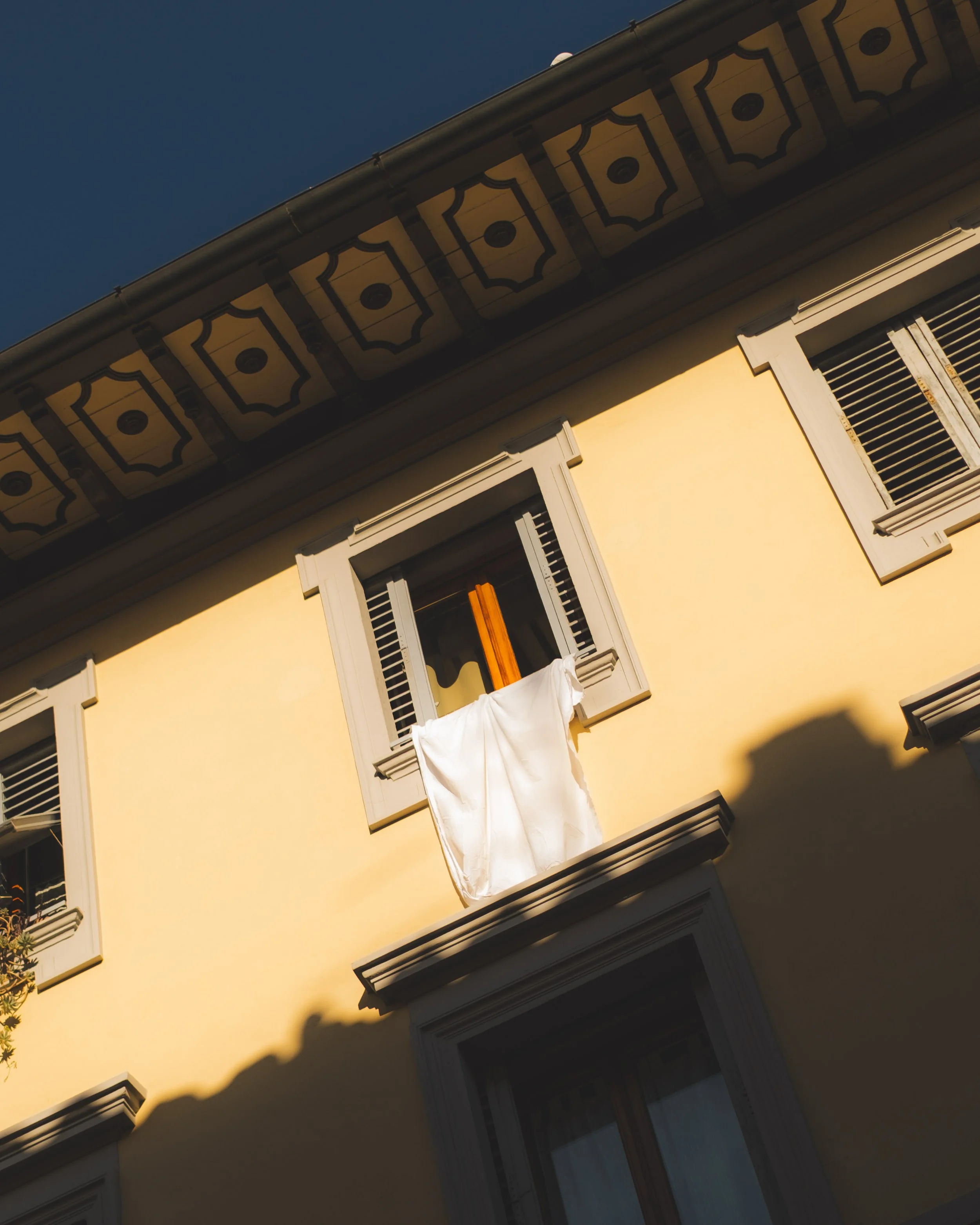 A yellow building with an open window and gray shutters, a white cloth hanging from the window, and decorative trim on the roof. Lionel Chu - Hotel & Travel Photographer