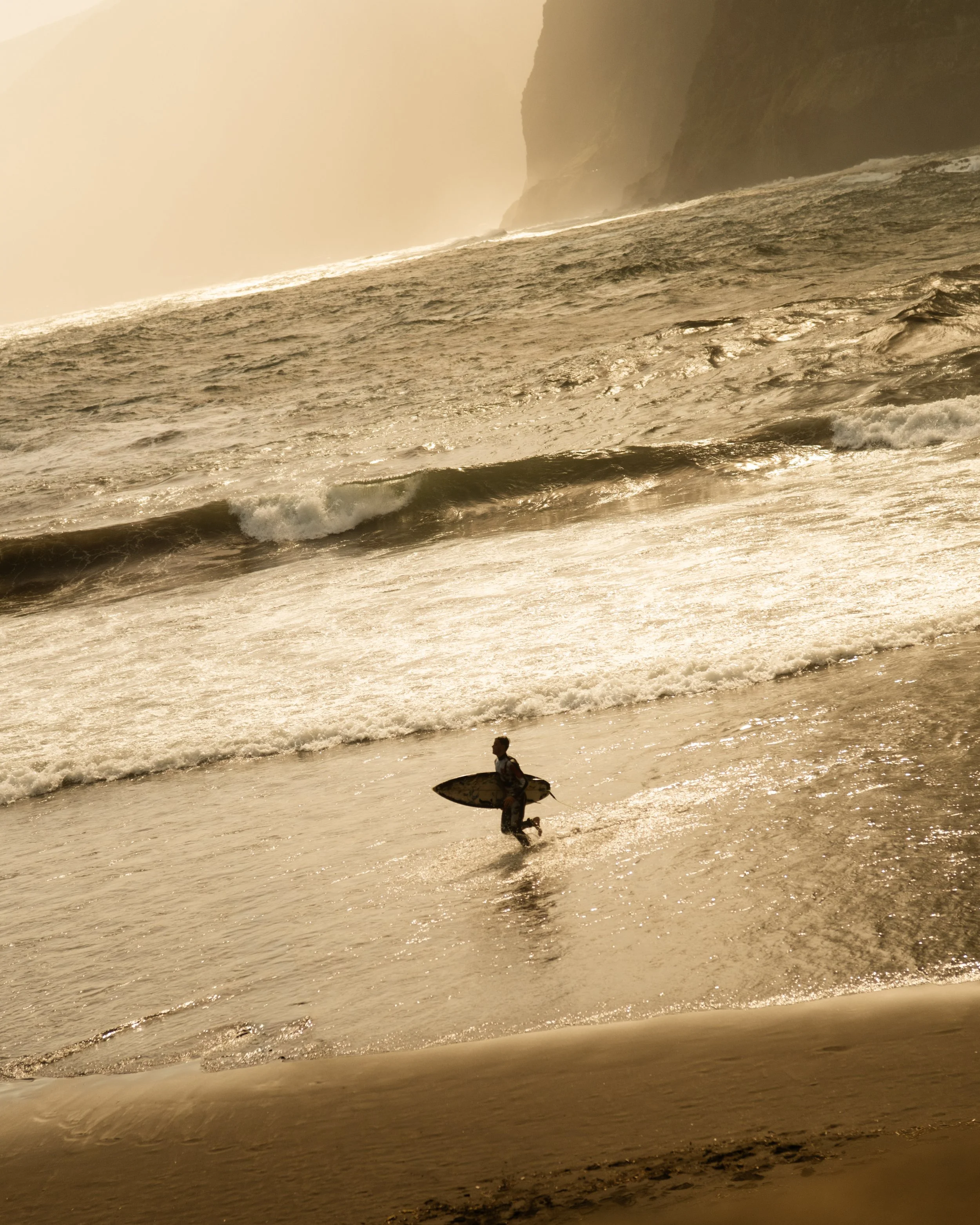 A person holding a surfboard and running on the beach near the ocean waves during sunset.