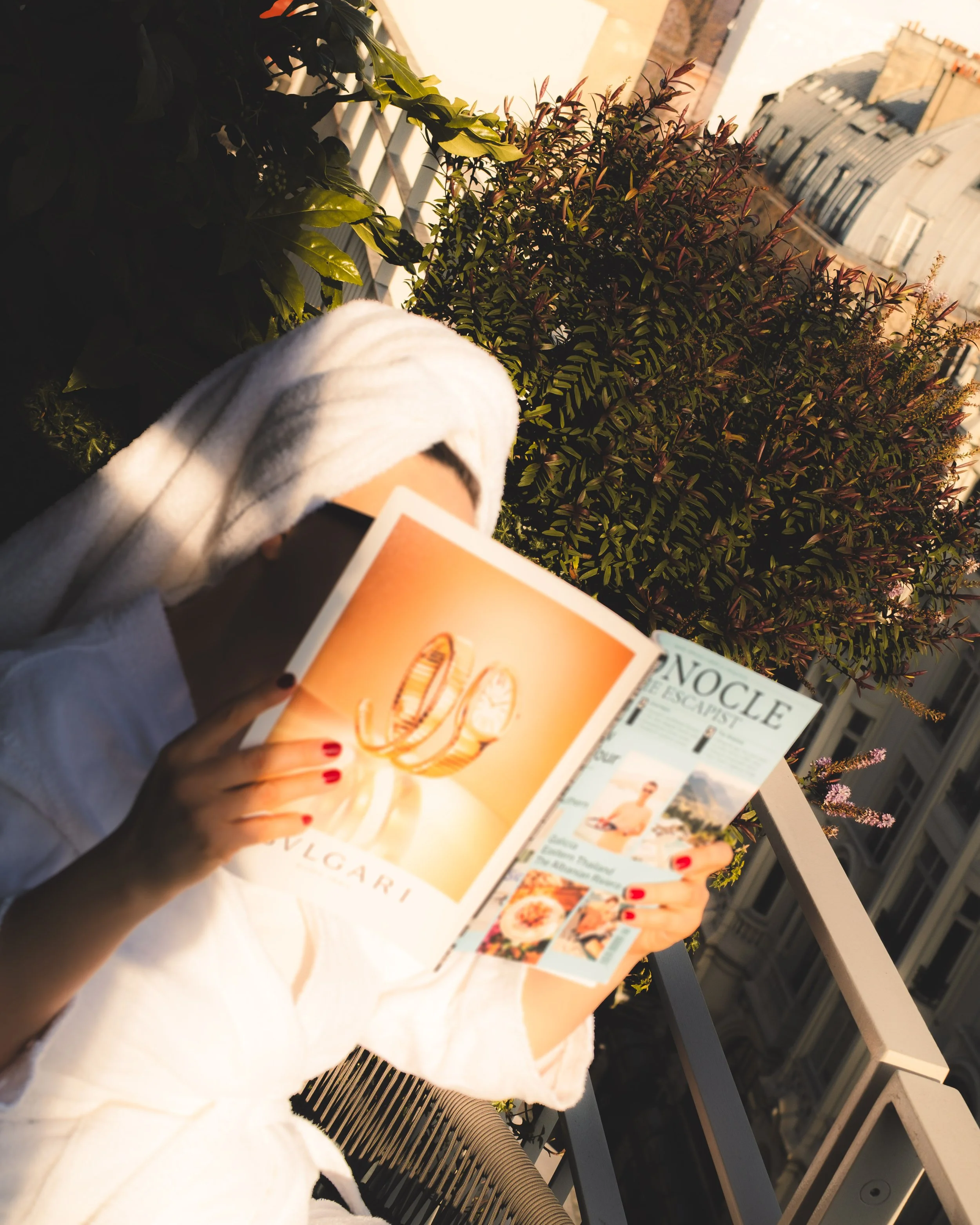 The image shows a person with red painted nails holding a magazine, standing on a balcony next to a table with some pamphlets and brochures, with plants and a building in the background during sunset.