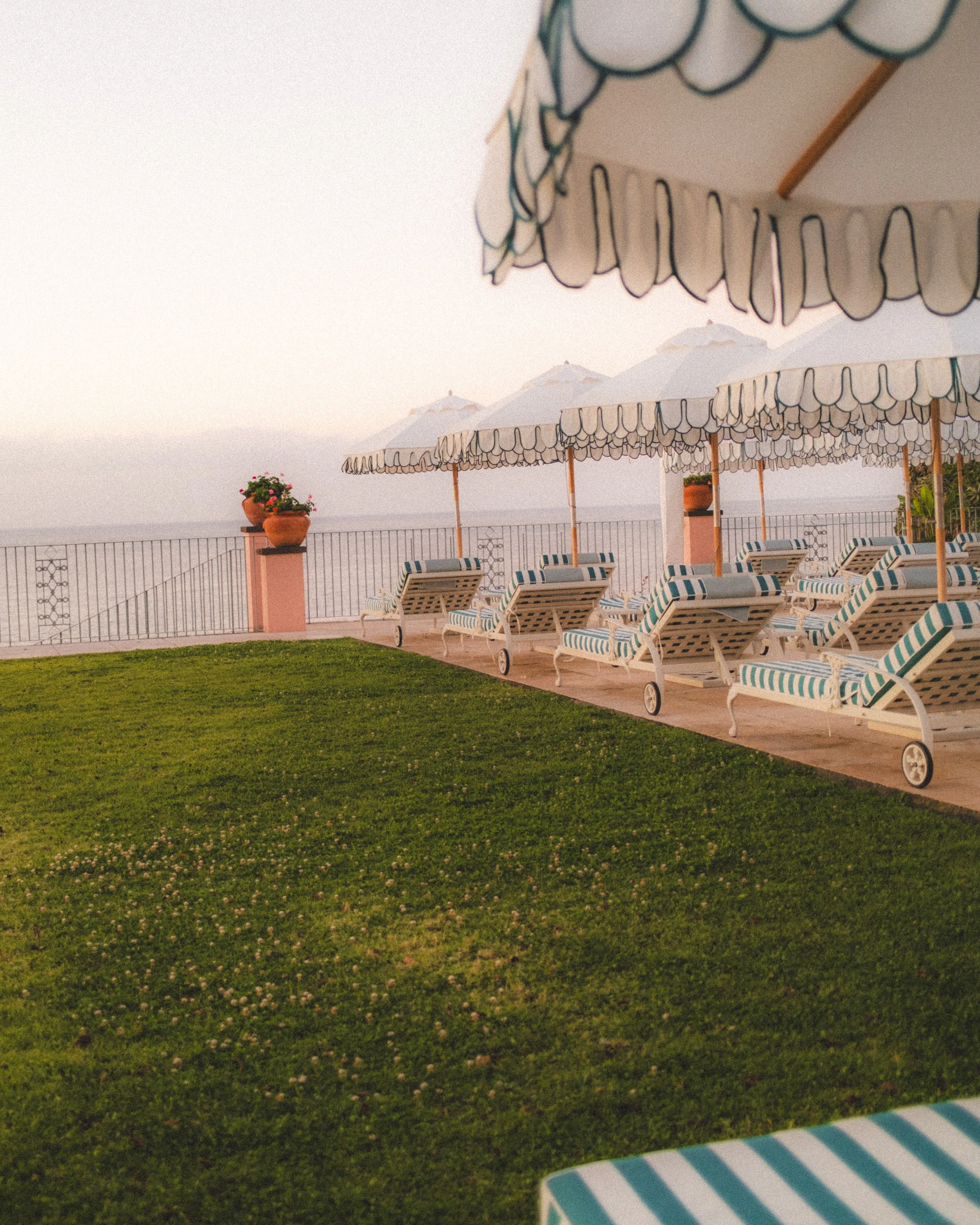 Beach scene with striped lounge chairs, large umbrellas, potted plants, a railing, and a distant view of the ocean and mountains at sunset.