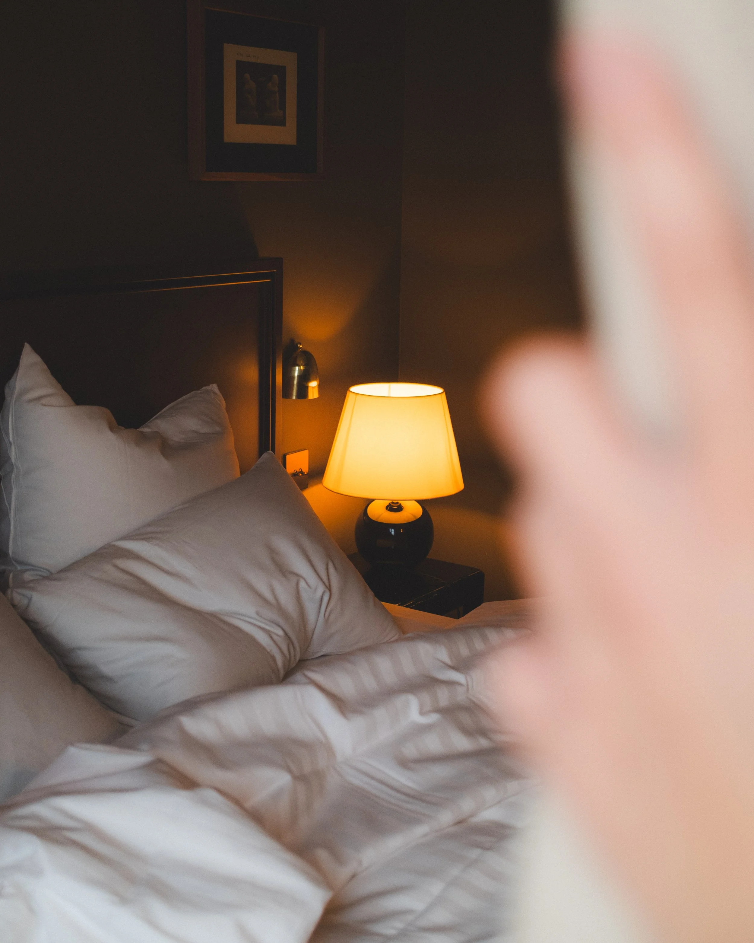 A bedroom scene with a bed, white pillows, a dark headboard, a bedside lamp, a table, and a framed picture on the wall.
