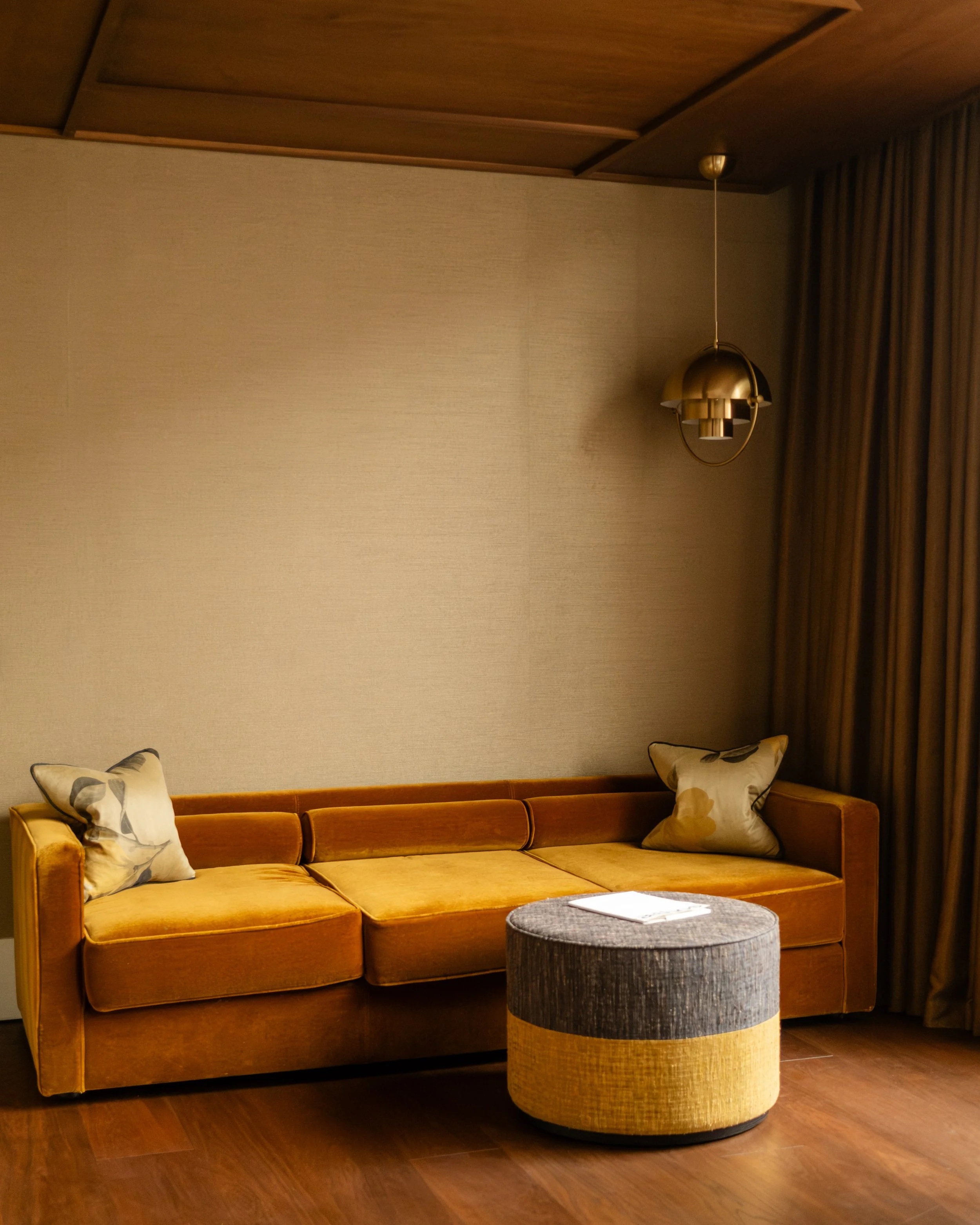 An hotel bedroom living room with a mustard yellow sofa, two patterned pillows, a round ottoman with a yellow and gray color block design, wooden flooring, a textured beige wall, and dark curtains, with a modern hanging brass light fixture.