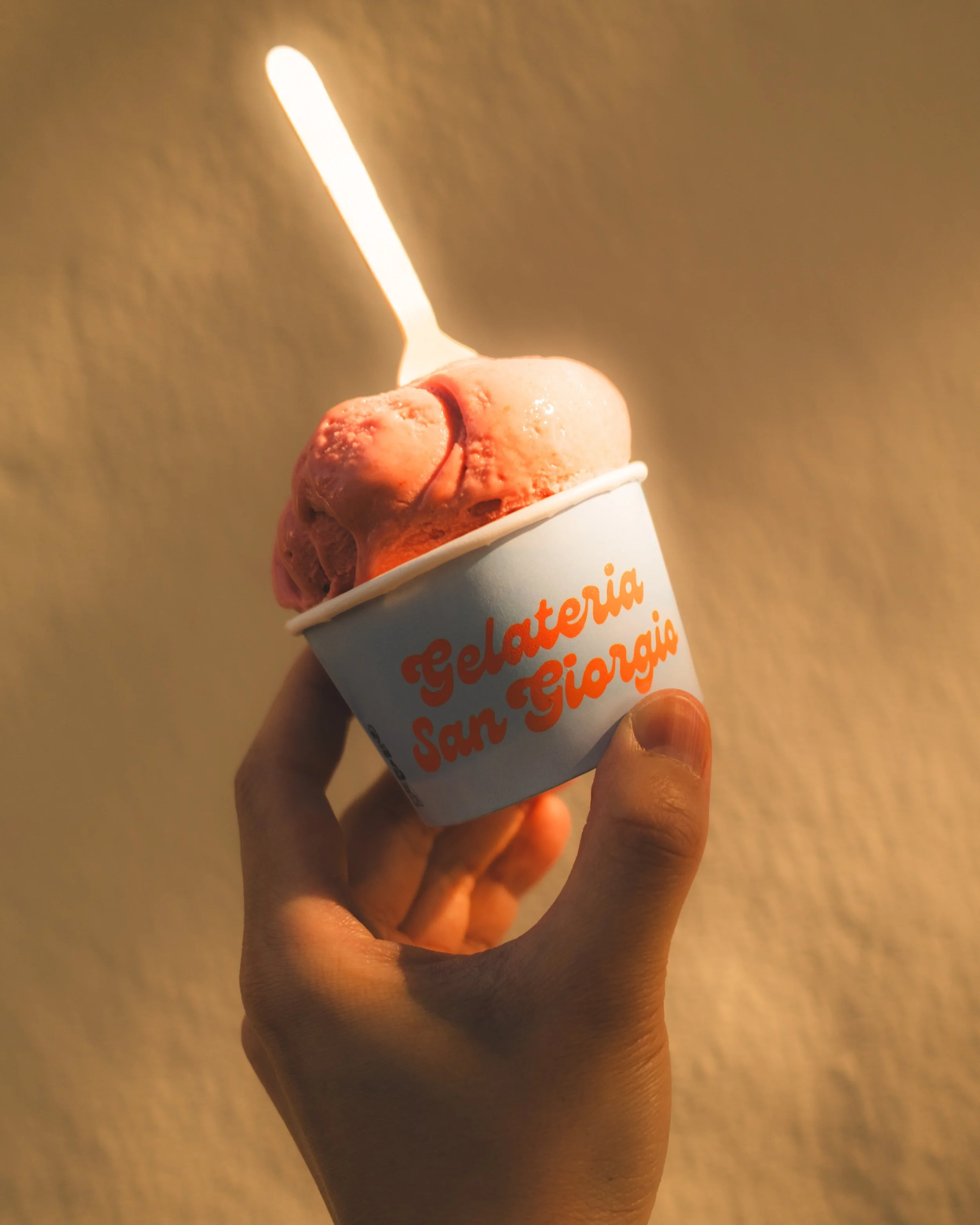 Person holding a cup of gelato with a matchstick candle on top. The gelato appears pink in color.