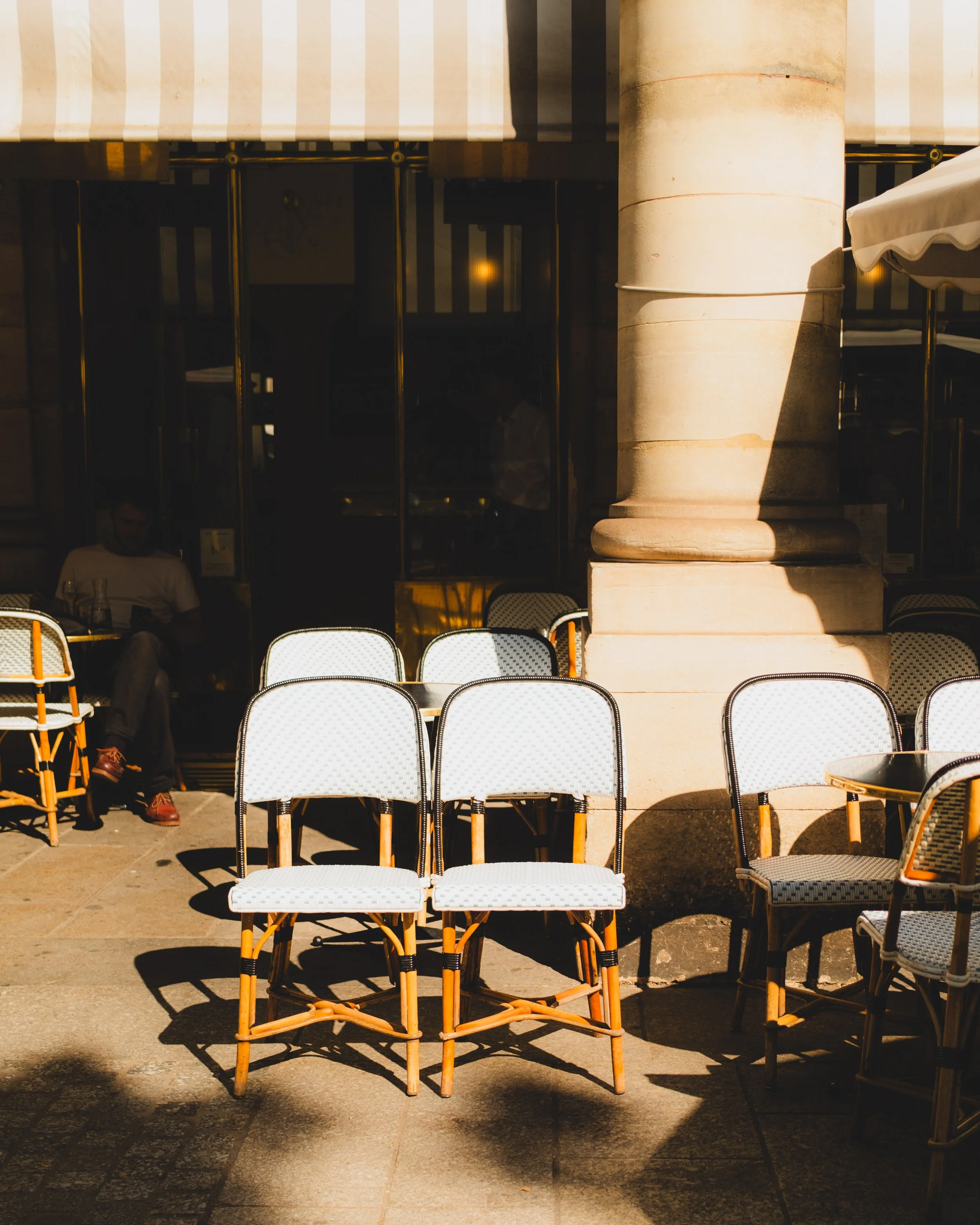 Empty outdoor cafe seating area with white and wicker chairs on a sunny day, shadowed by a large column and striped awning. Lionel Chu - Hotel & Travel Photographer