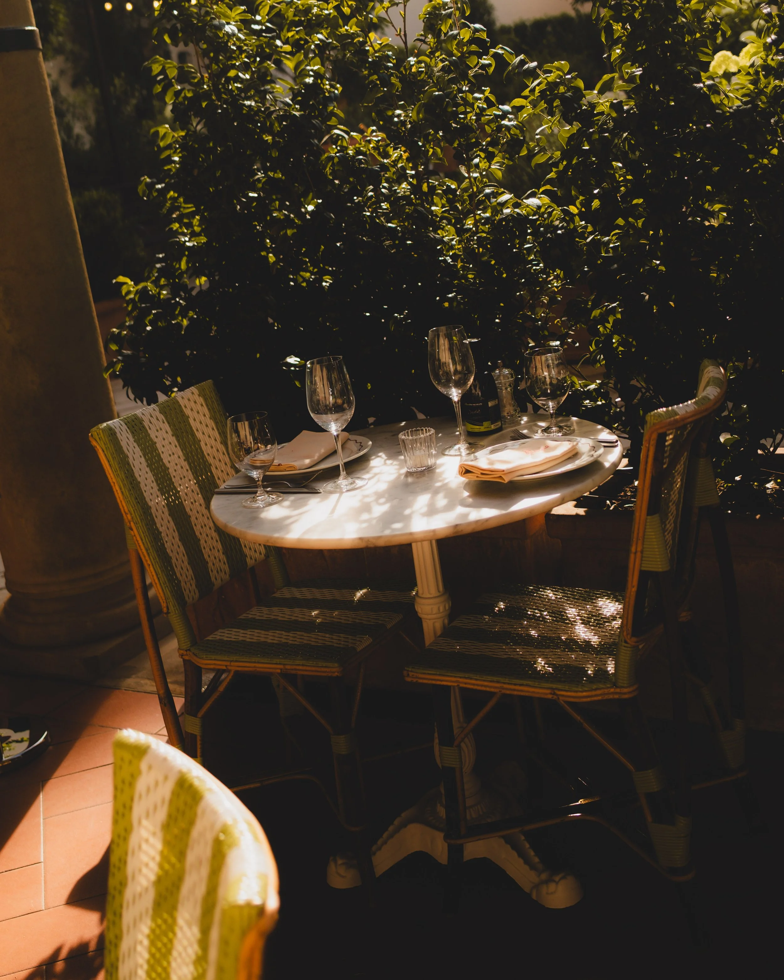 An outdoor dining table set for a meal with wine glasses, plates, and napkins, surrounded by chairs, with a lush green hedge in the background and warm evening sunlight.