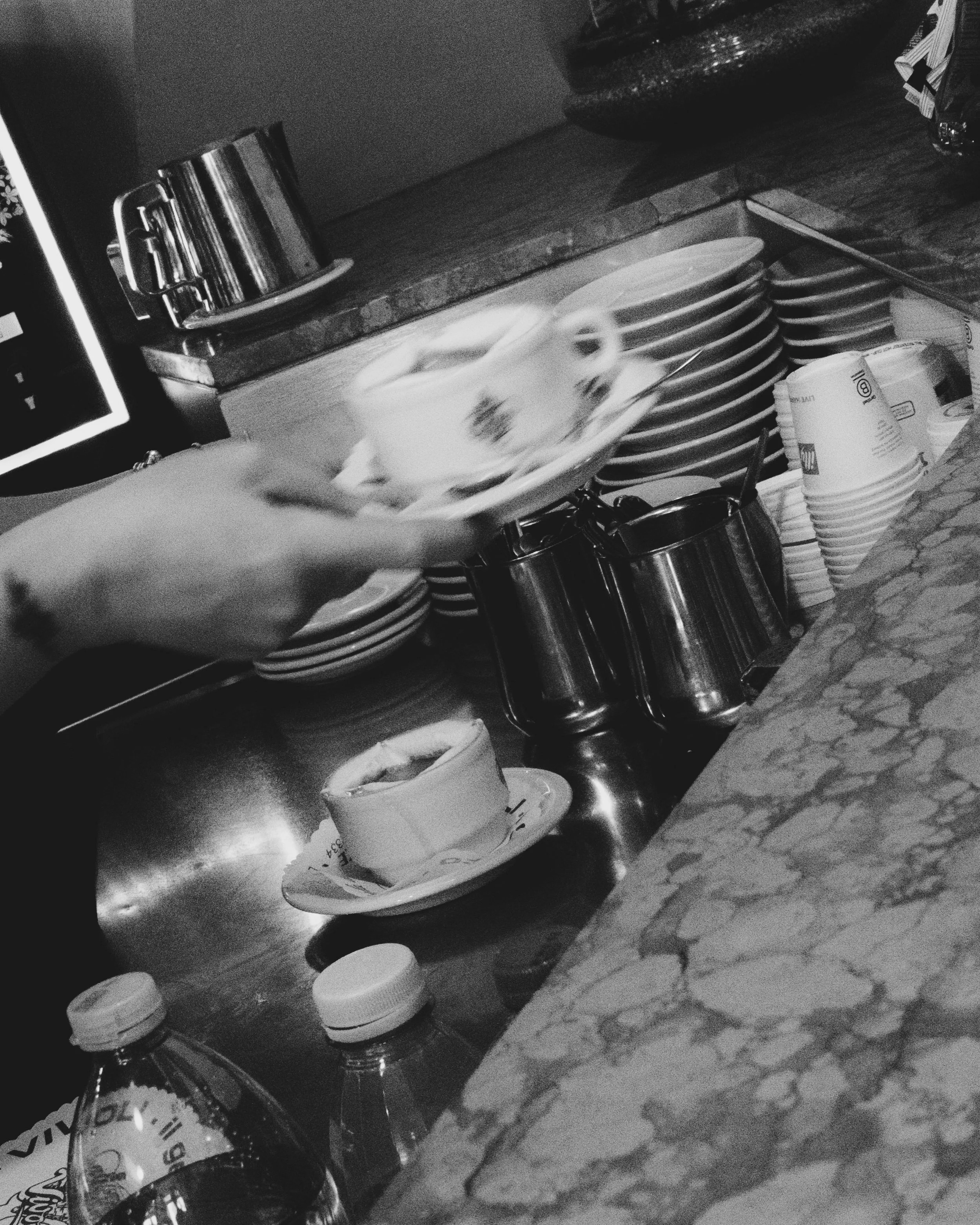 A person serving a cup of coffee or tea in a cafe with stacked plates, cups, and water bottles on a marble counter. Lionel Chu - Hotel & Travel Photographer