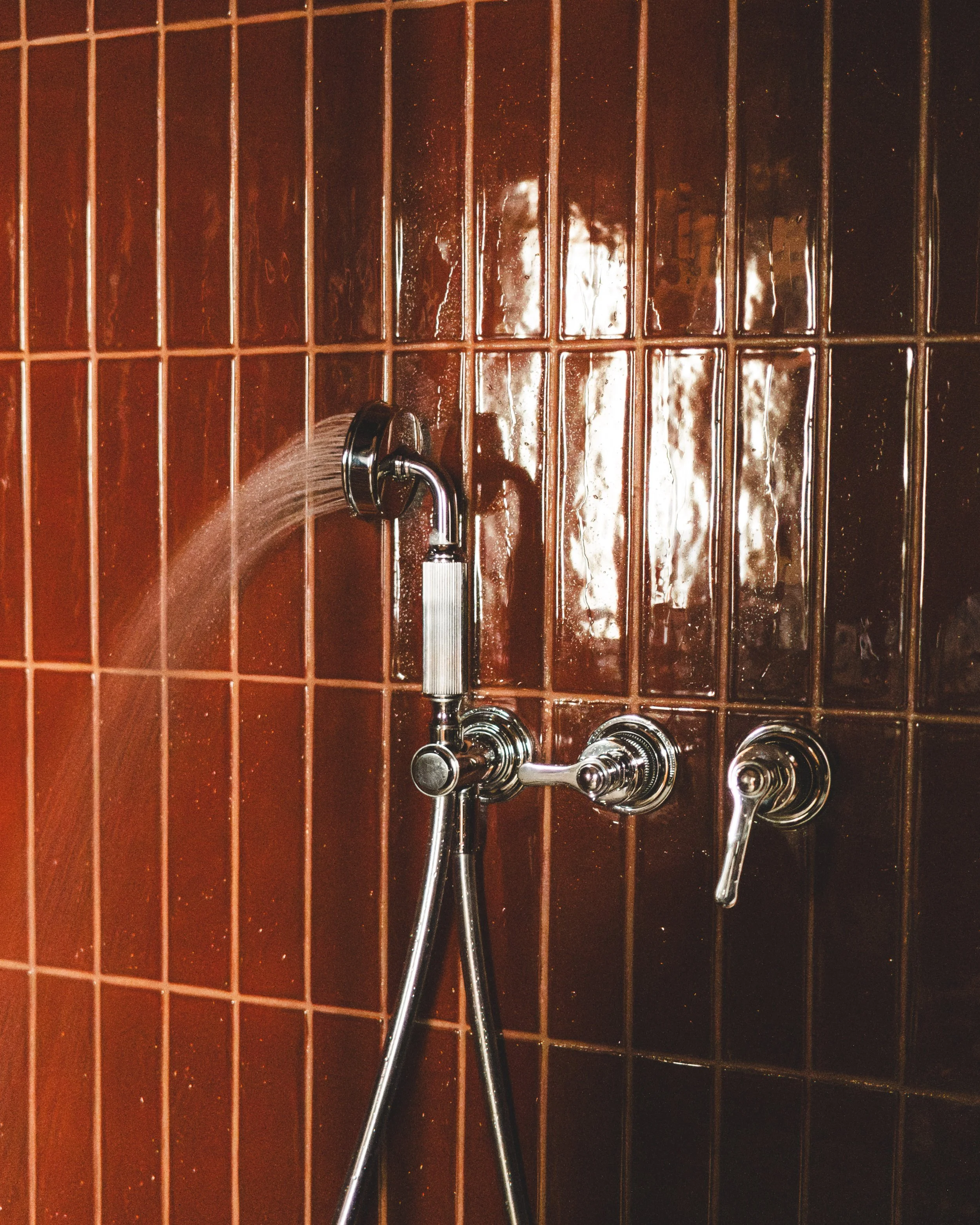 A shower with brown tile walls, a chrome showerhead, a handheld shower wand, and a water faucet. Water is flowing from the showerhead.