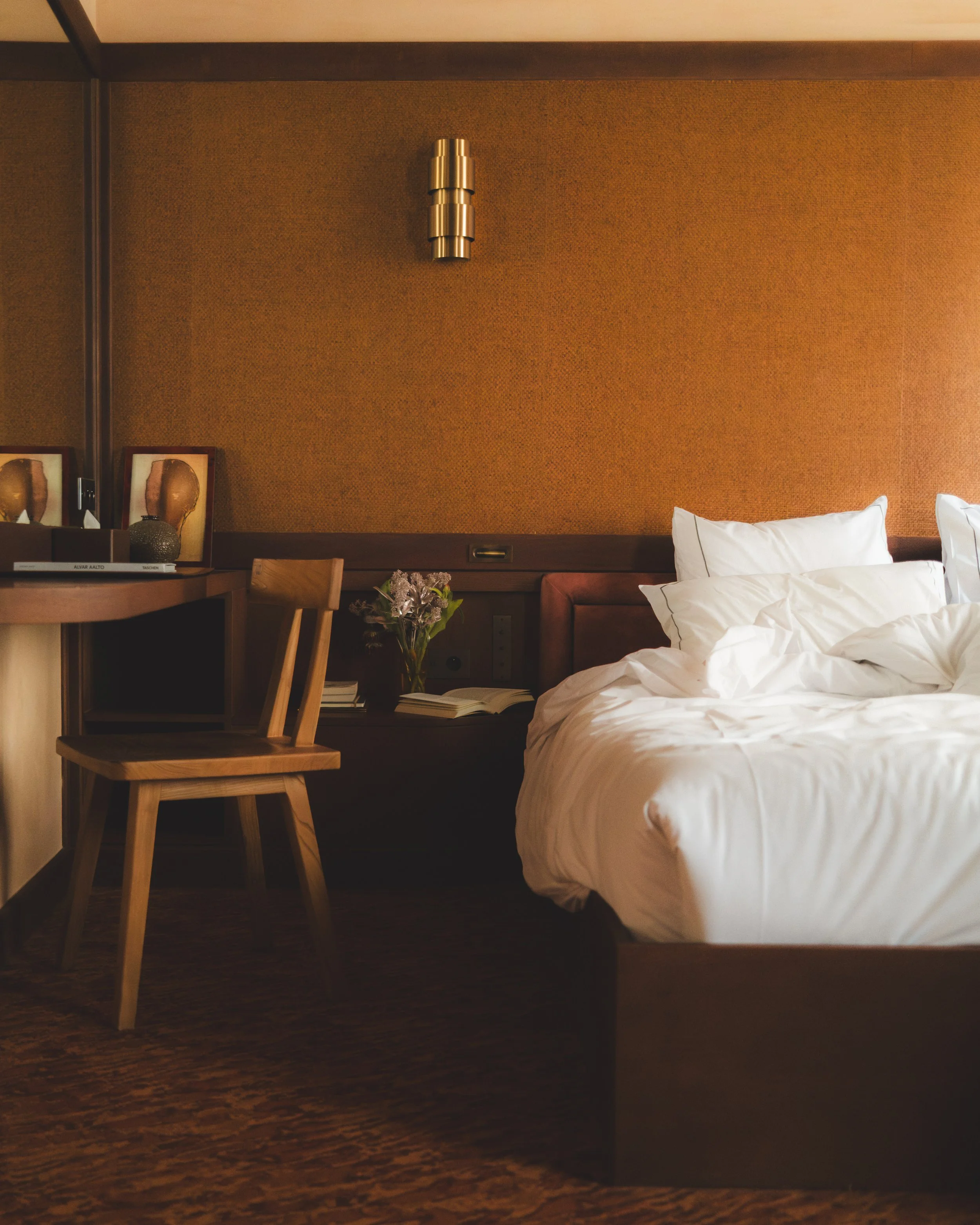 A bedroom corner with a bed, nightstand, and decorative pillows, featuring a textured, burnt orange wall with modern metallic wall art and a small wooden dining table with a chair.