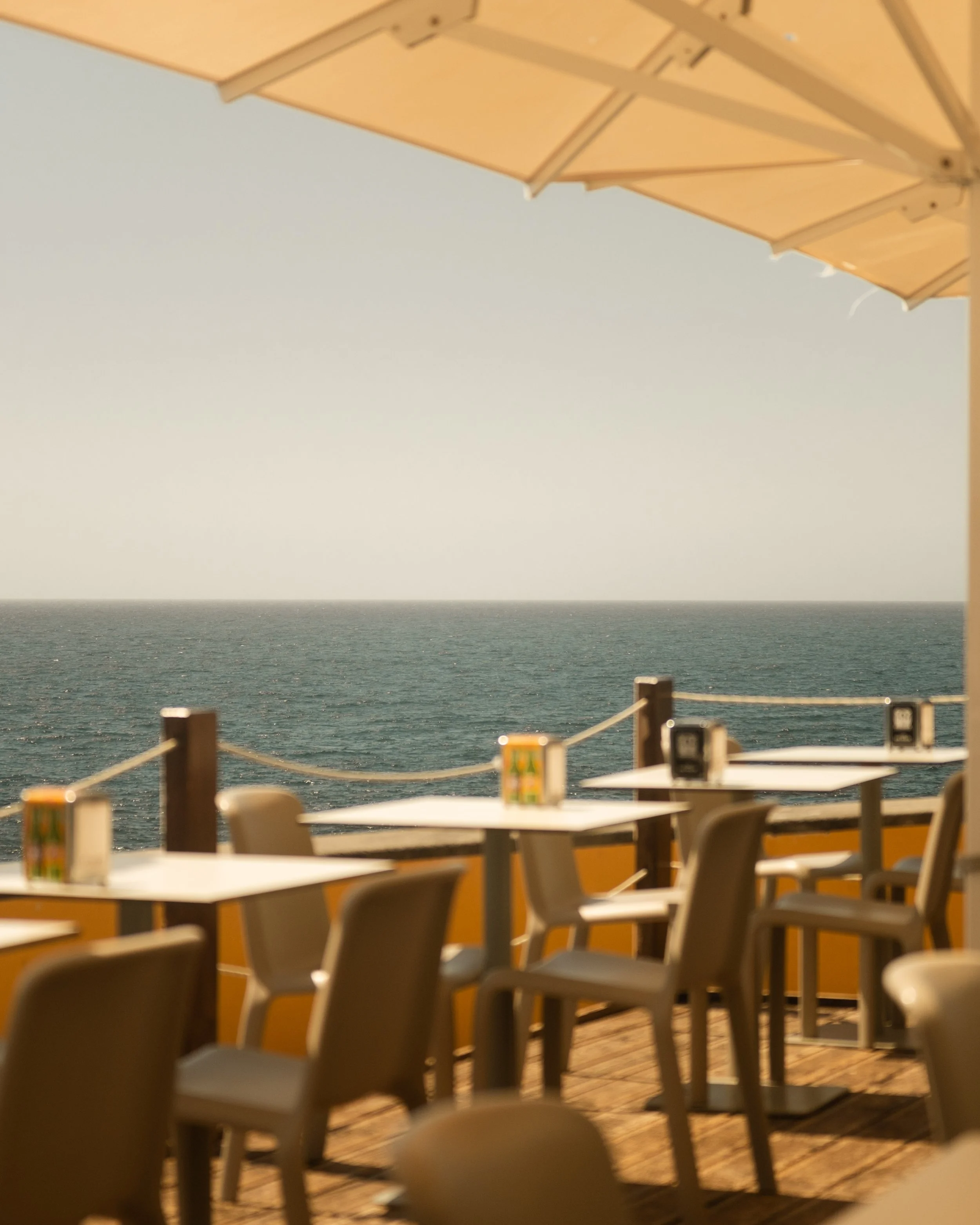 Blank tables and chairs outside on a deck or patio overlooking the ocean with a railing. The sky is clear and sunny.