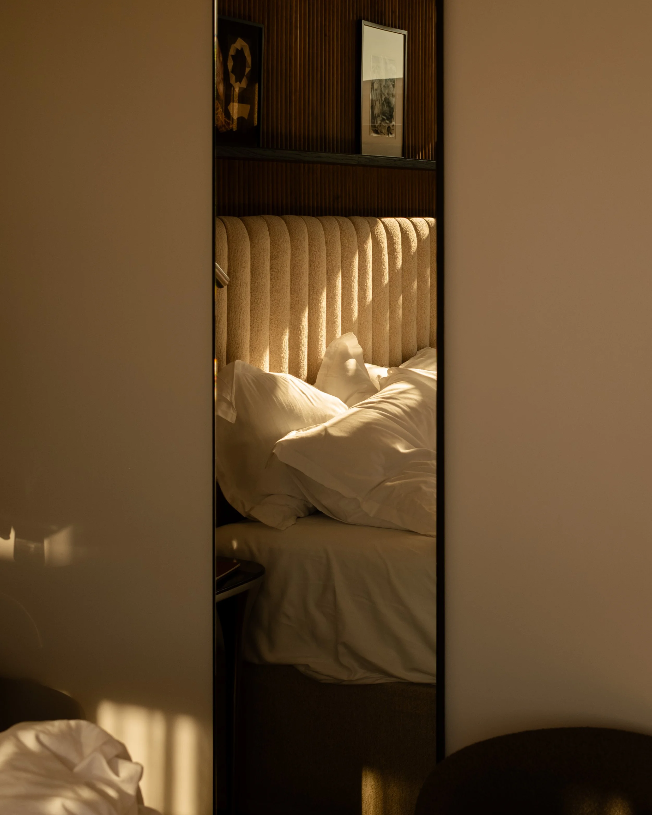 A hotel room with a bed seen through a mirrored closet door; the bed has white sheets and pillows, and a beige headboard with a wooden wall and framed artwork behind it. Lionel Chu - Hotel & Travel Photographer