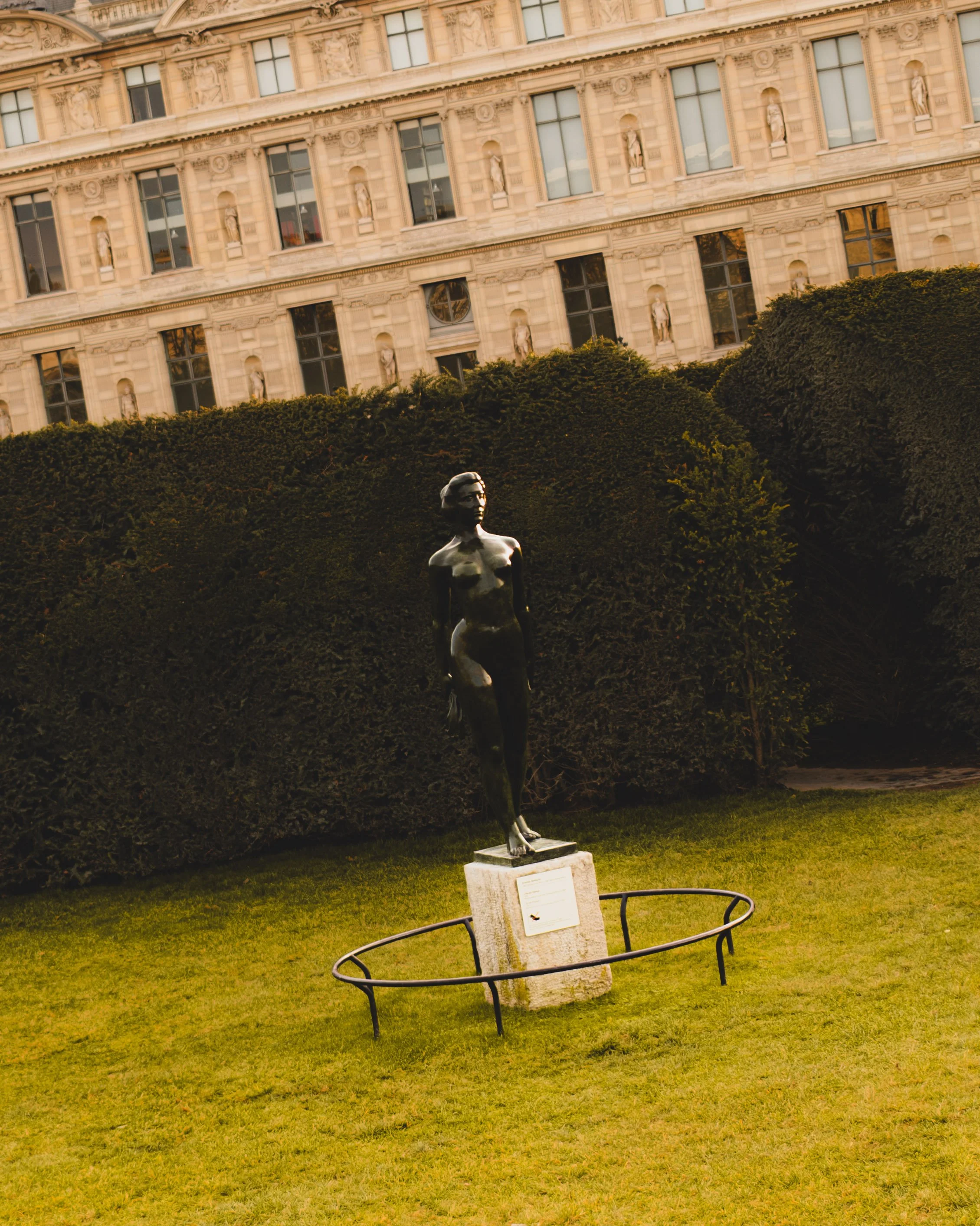Bronze sculpture of a woman standing outdoors in the Tuileries Gardent on a stone pedestal, surrounded by a low metal railing, with a hedge and the Louvre Museum in the background. Lionel Chu - Hotel & Travel Photographer