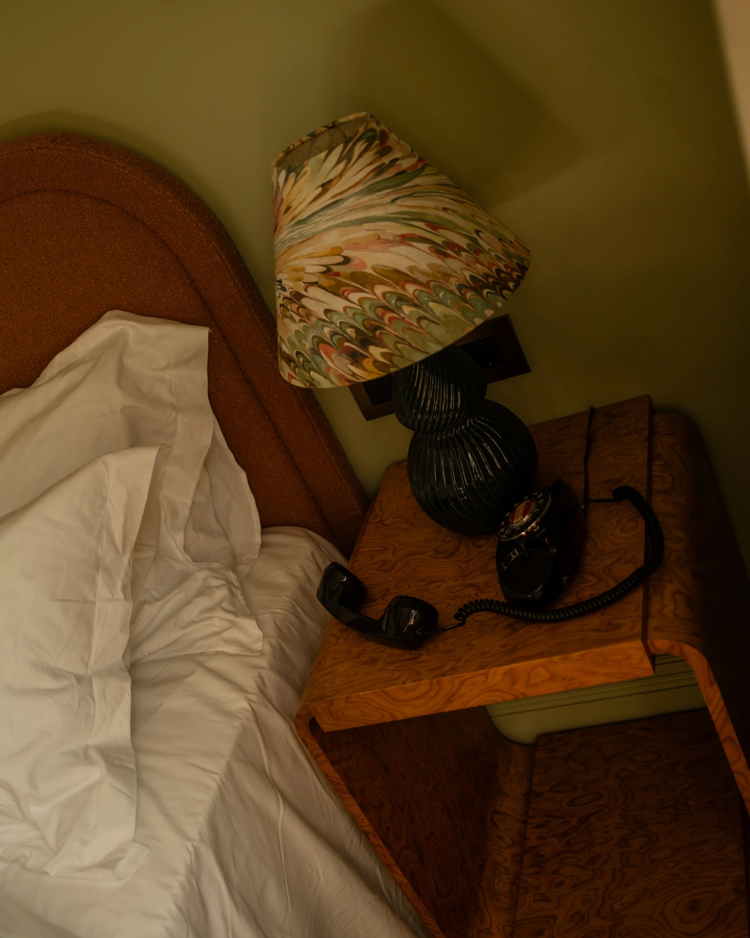 A bedroom with a bed, wooden nightstand, vintage lamp with a multicolored patterned shade, and a black rotary phone. Lionel Chu - Hotel & Travel Photography
