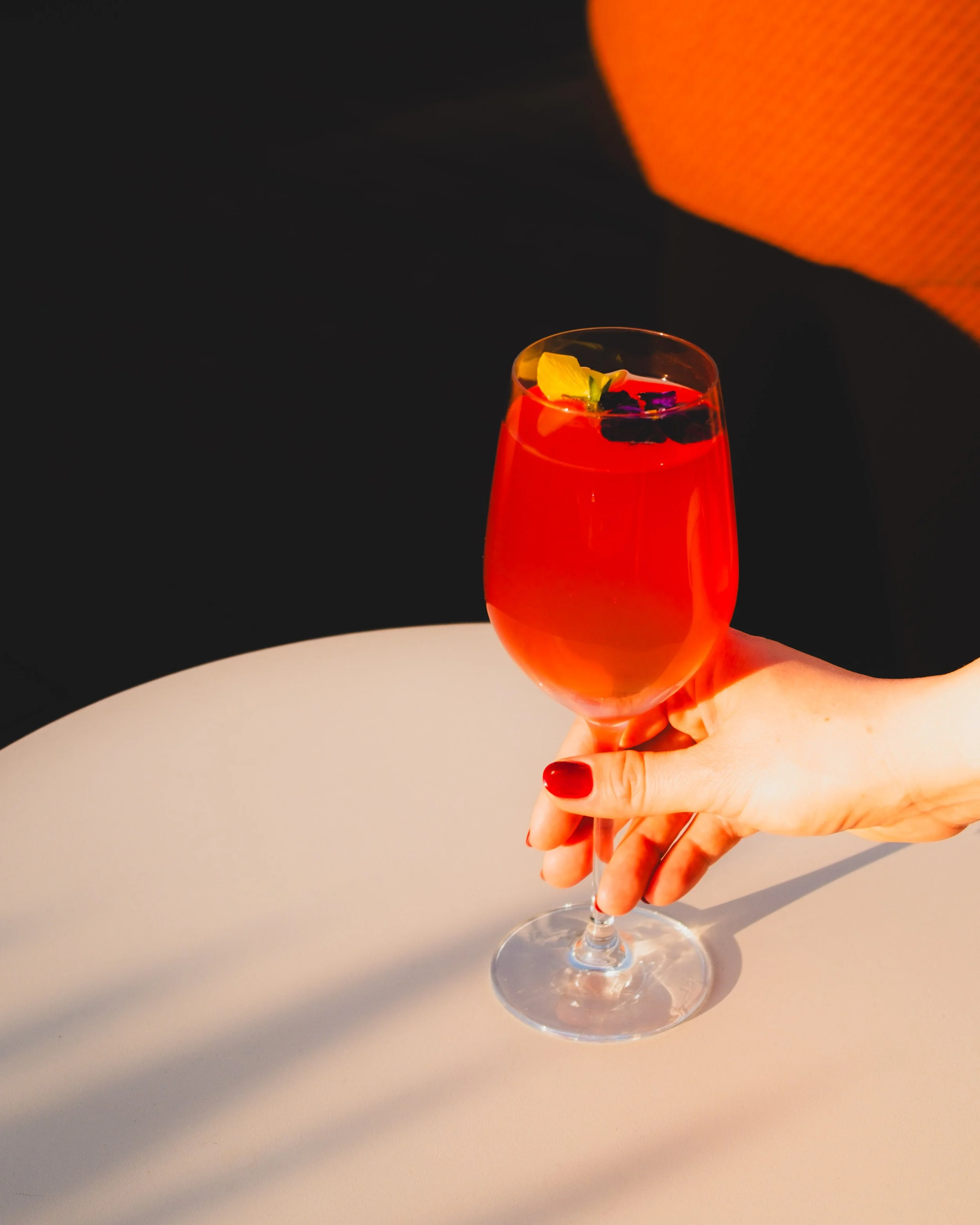 A hand with red painted nails holding a glass of red cocktail with garnishes on top against a background of black and orange fabrics.