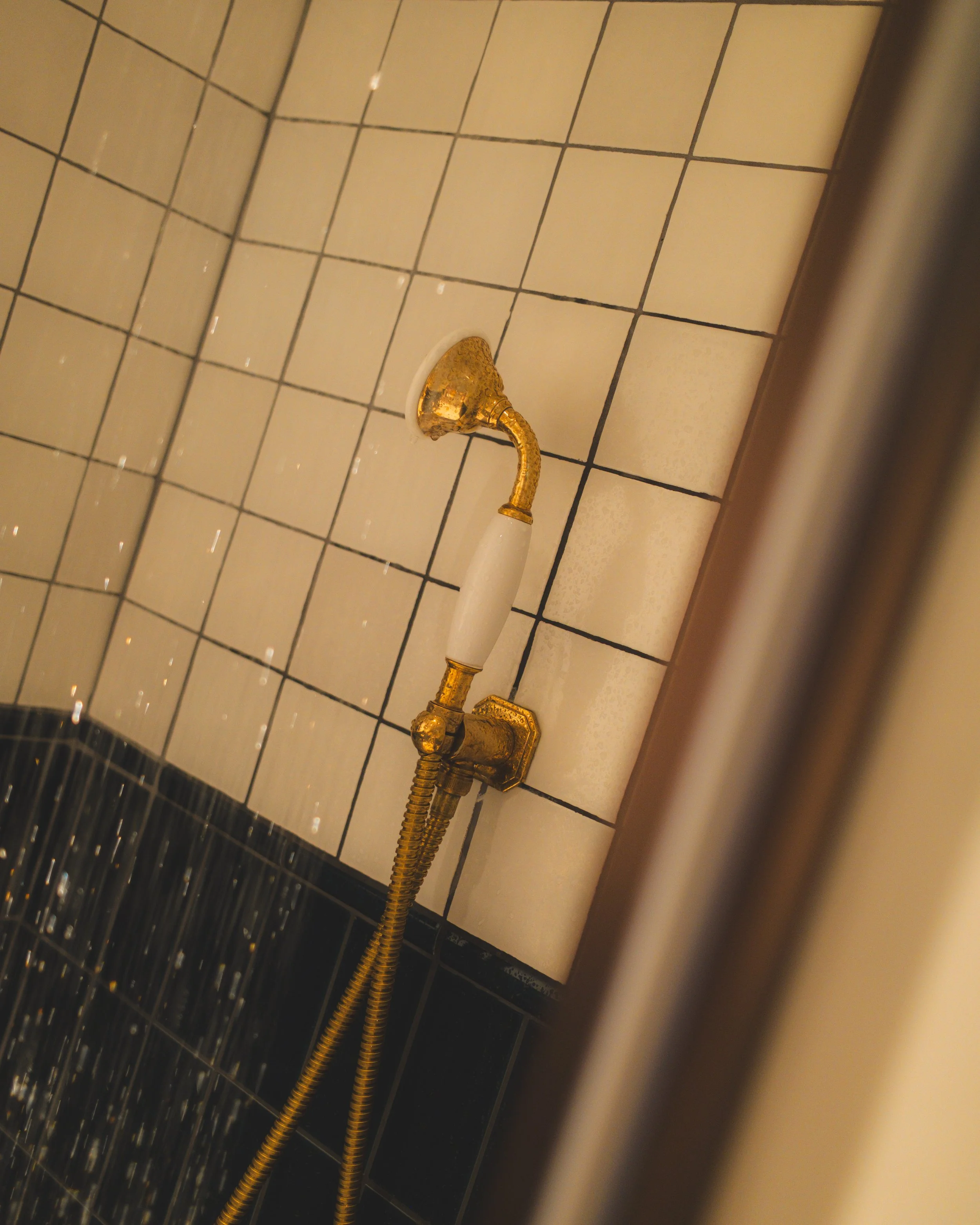 View of a gold-colored faucet attached to a wall with white tiled surface, and black tiled flooring beneath.