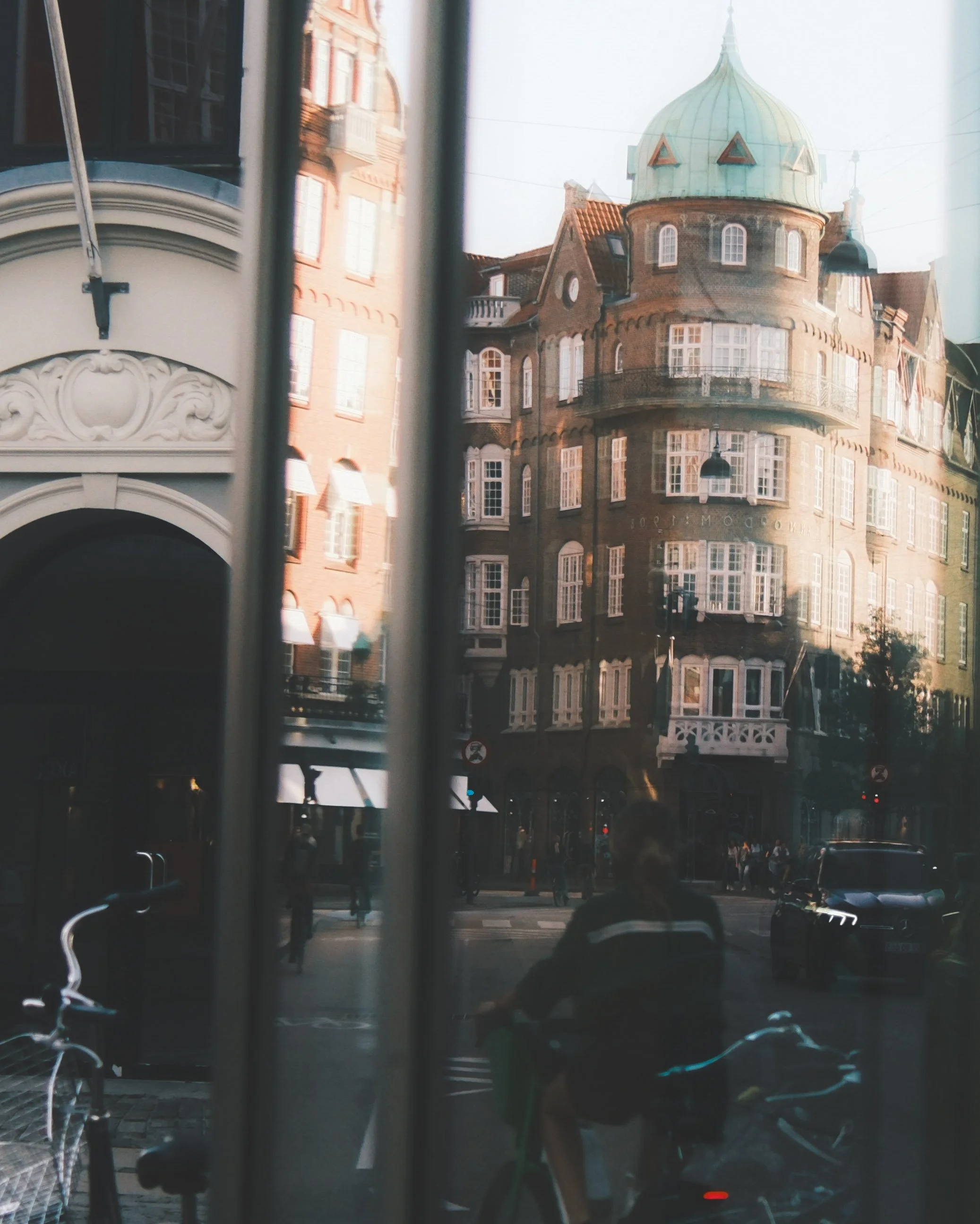 View through a window showing historic brick buildings with ornate architectural details, a rounded tower with a green domed roof, and city street with pedestrians and traffic.
