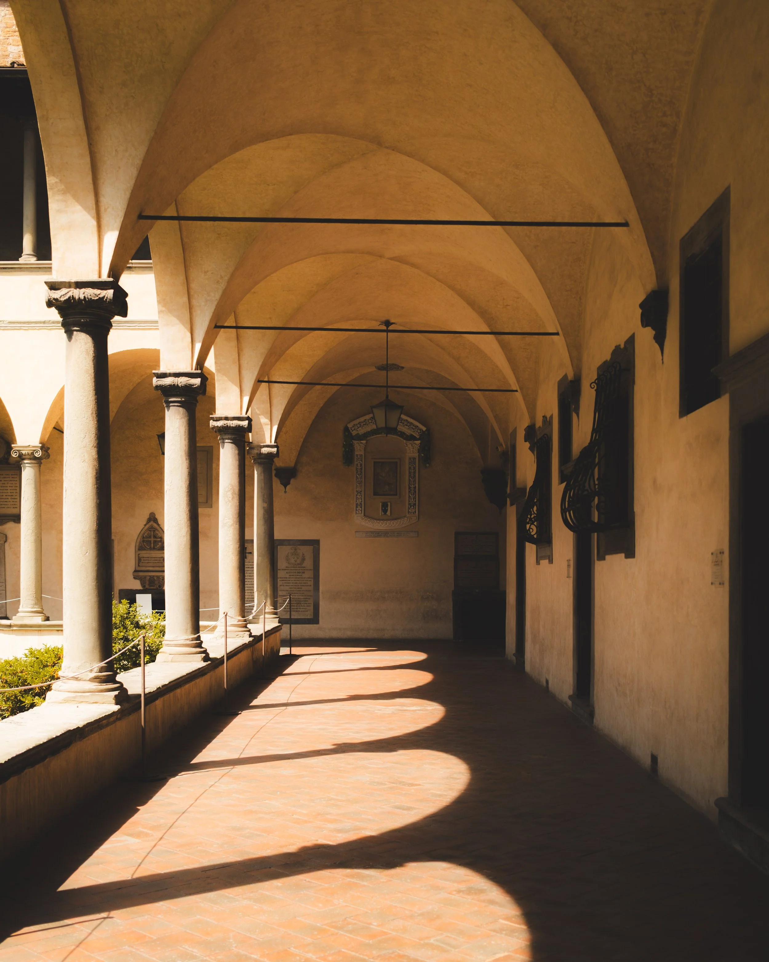 Arcade with tall columns, vaulted ceilings, and hanging lights illuminated by sunlight creating shadows on the stone floor.