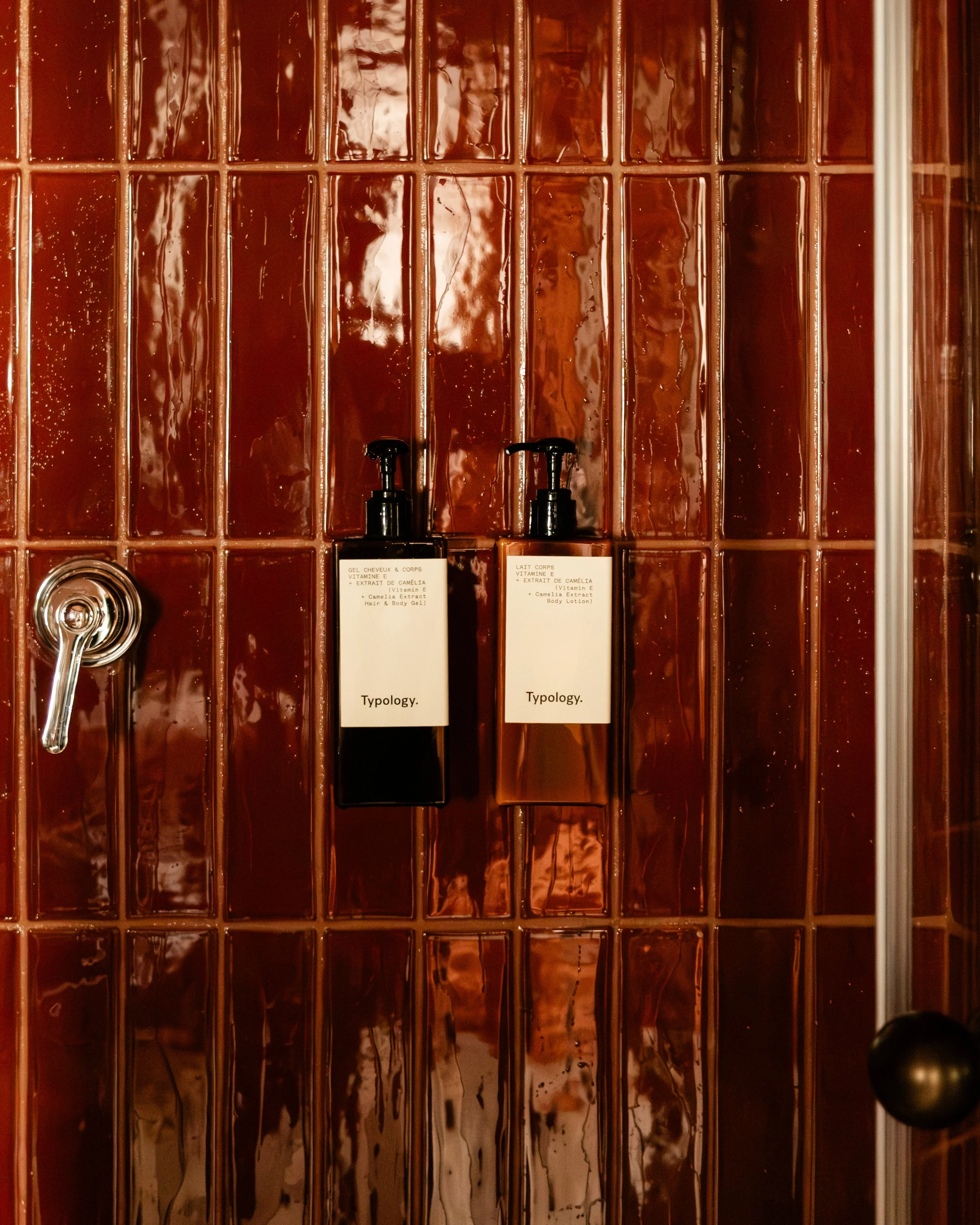 Two bottles of body lotion mounted on a glossy reddish-brown tiled wall, of an hotel bathroom. Lionel Chu - Hotel & Travel Photography