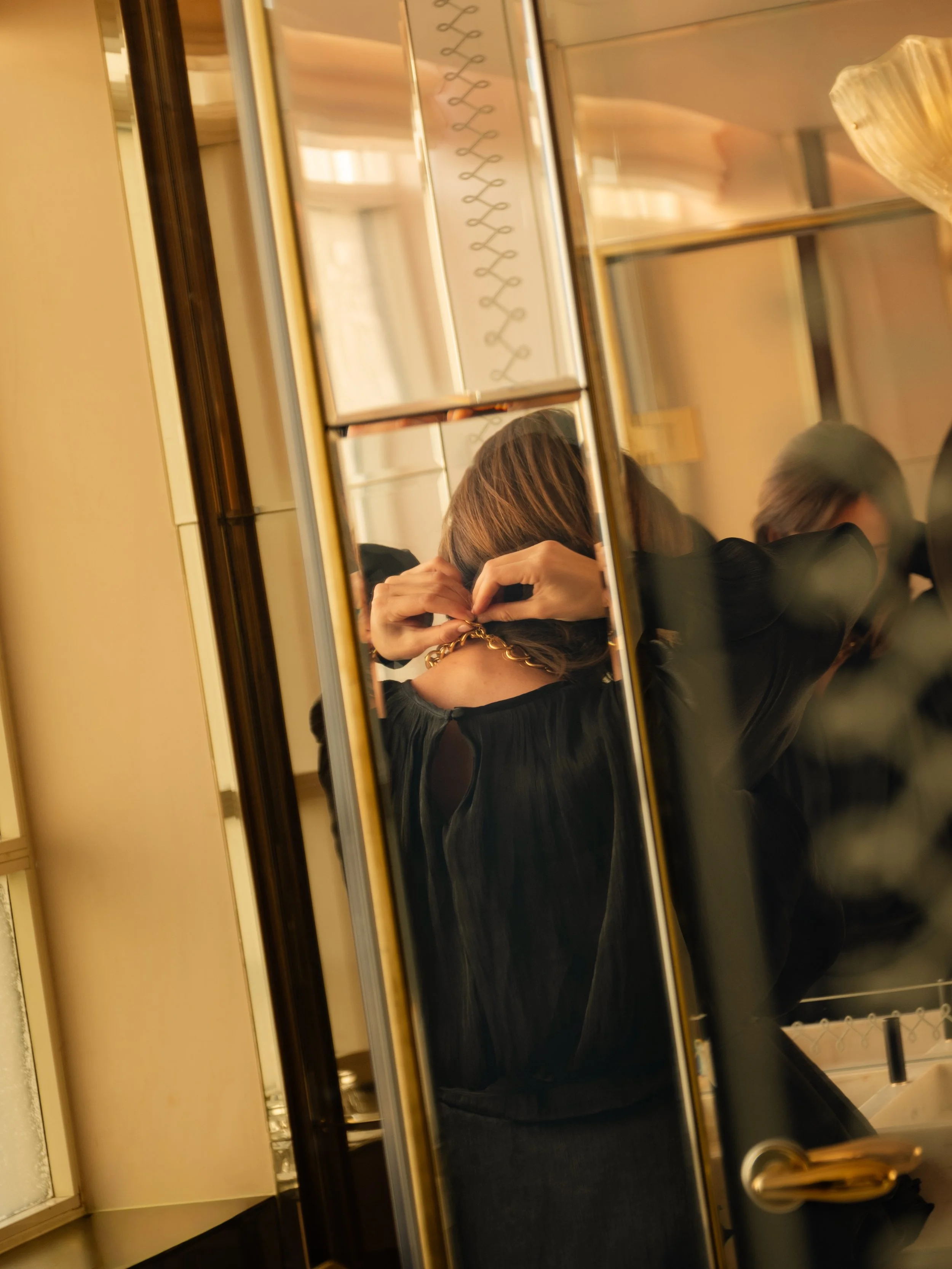 A woman in black is seen from behind, adjusting a gold chain necklace in front of a mirror in a bathroom, with her reflection visible. Lionel Chu - Hotel & Travel Photography