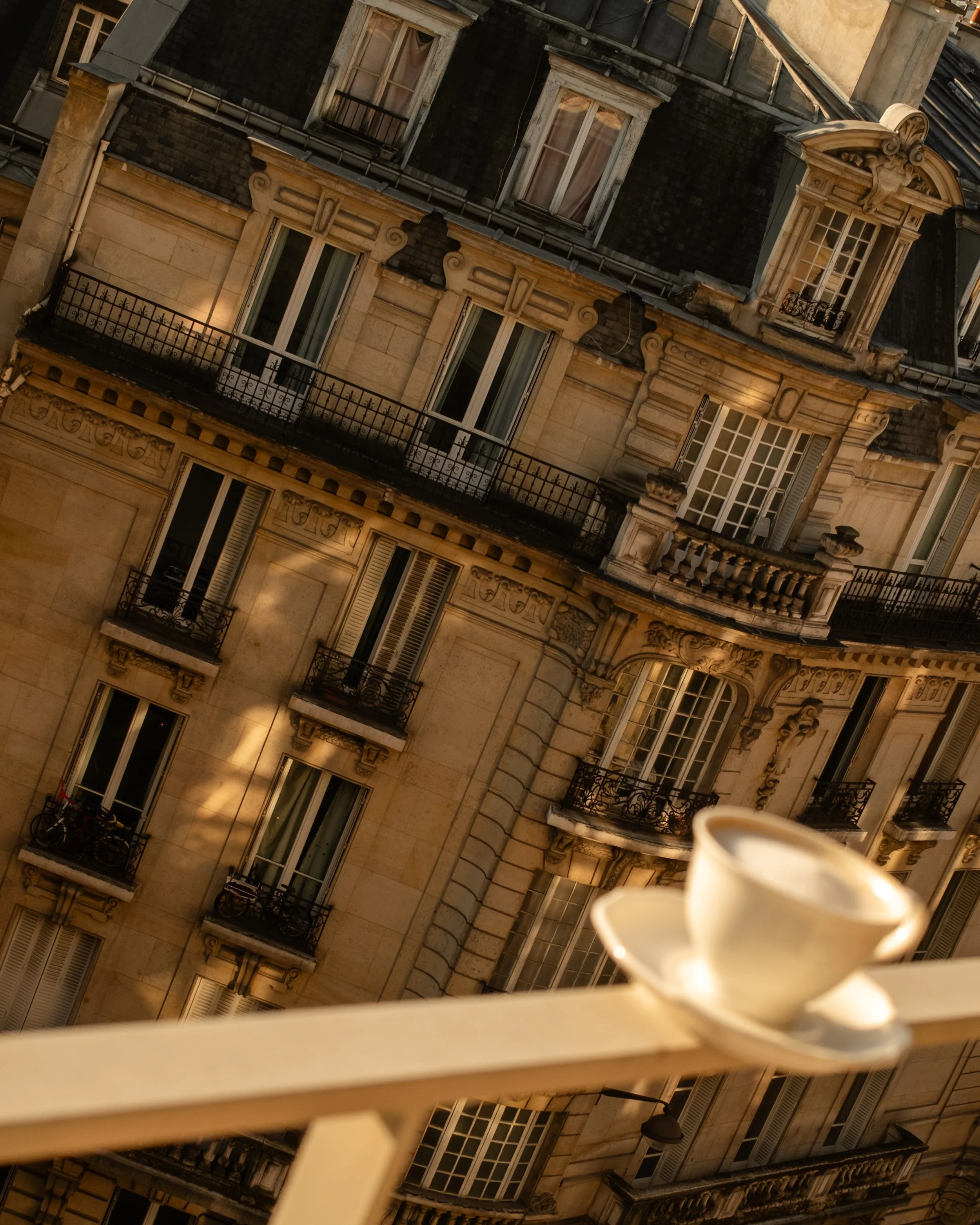 A white coffee cup on a saucer resting on a balcony railing, with a multi-story beige building with ornate windows and balconies in the background. Lionel Chu - Hotel & Travel Photographer