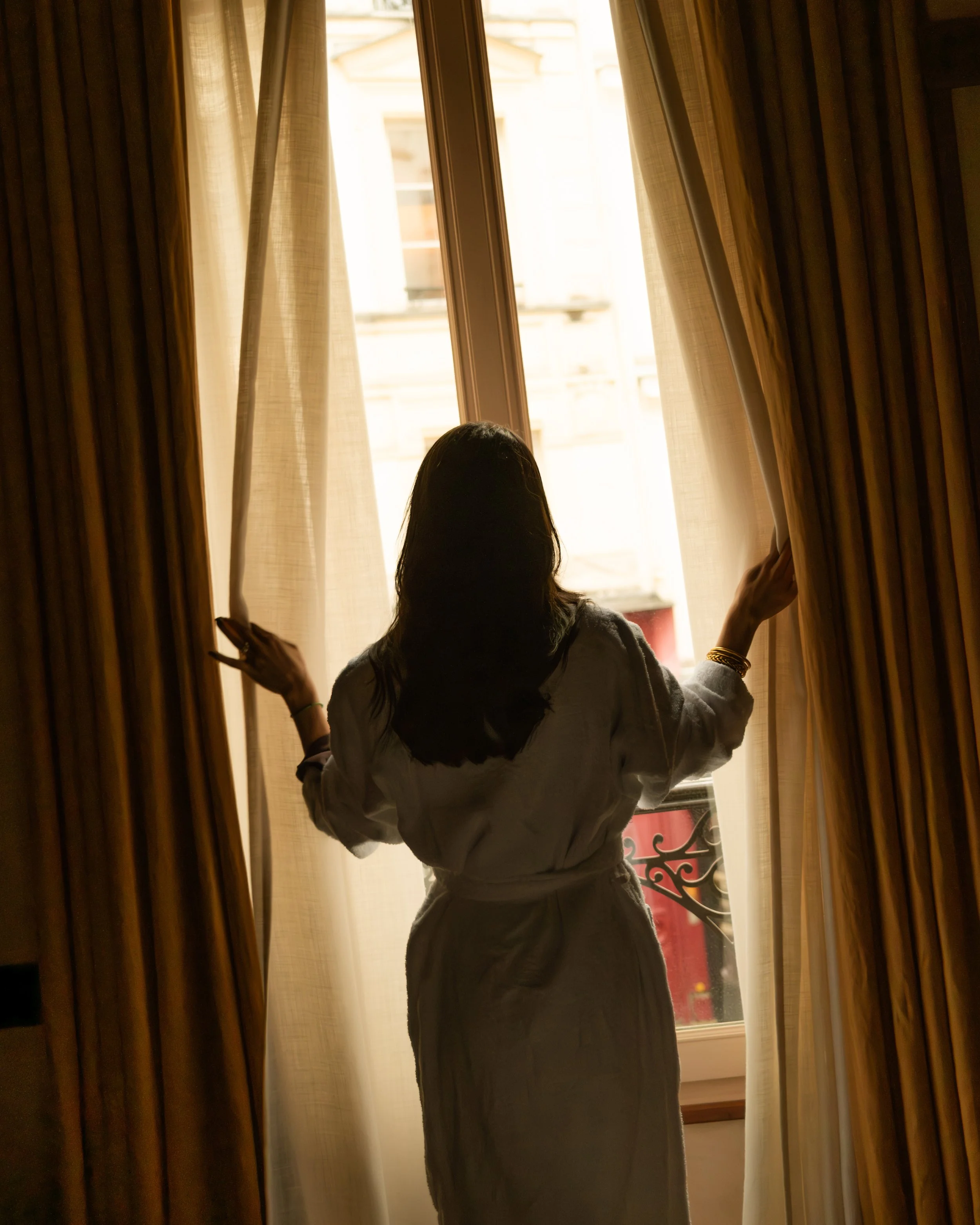 A woman with dark hair is standing by a window, opening curtains and looking outside. The scene is backlit, creating a silhouette effect. Lionel Chu - Hotel & Travel Photography