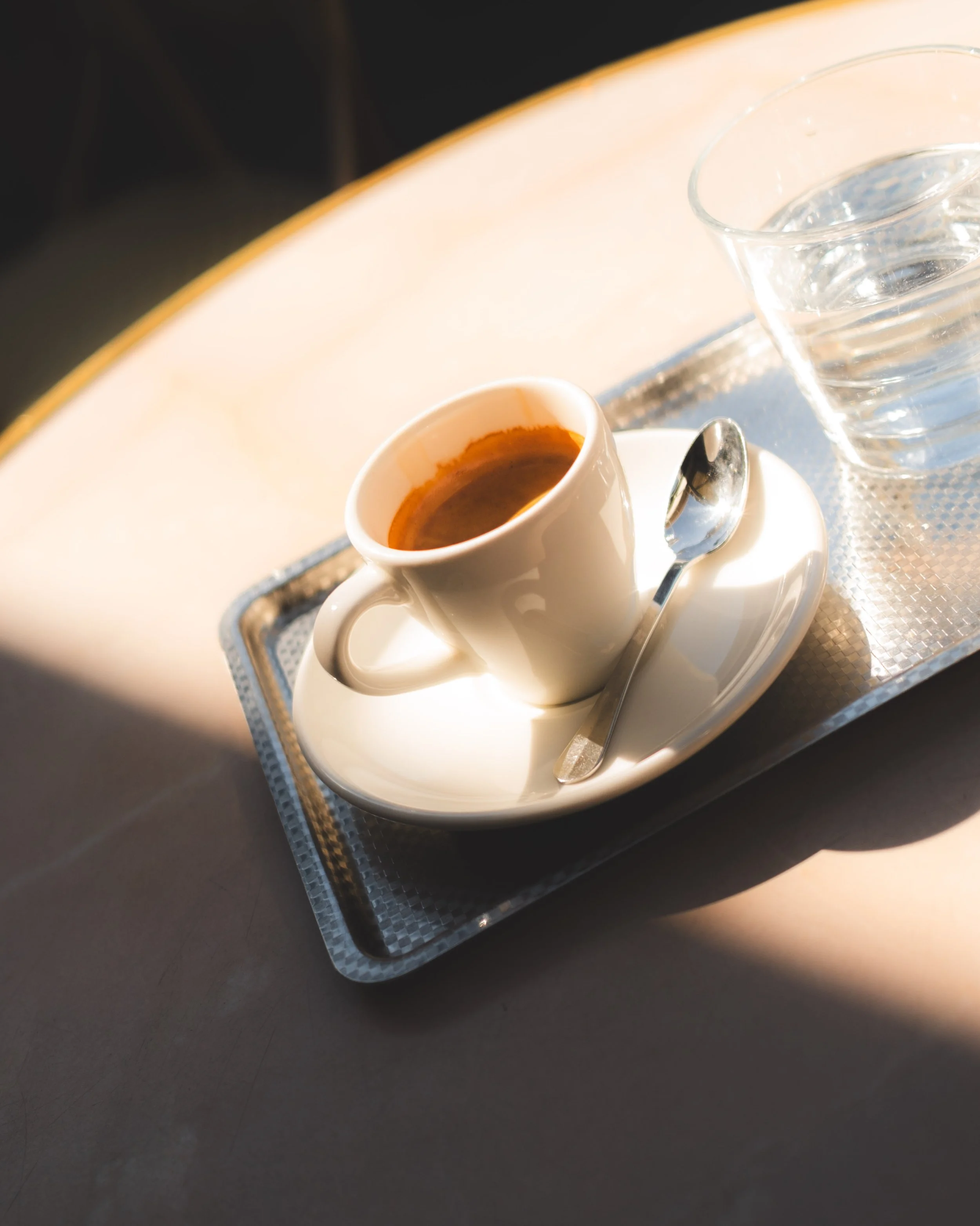 A tray holding a cup of black coffee, a glass of water, and a teaspoon, placed on a reflective surface with a curved edge in the background.