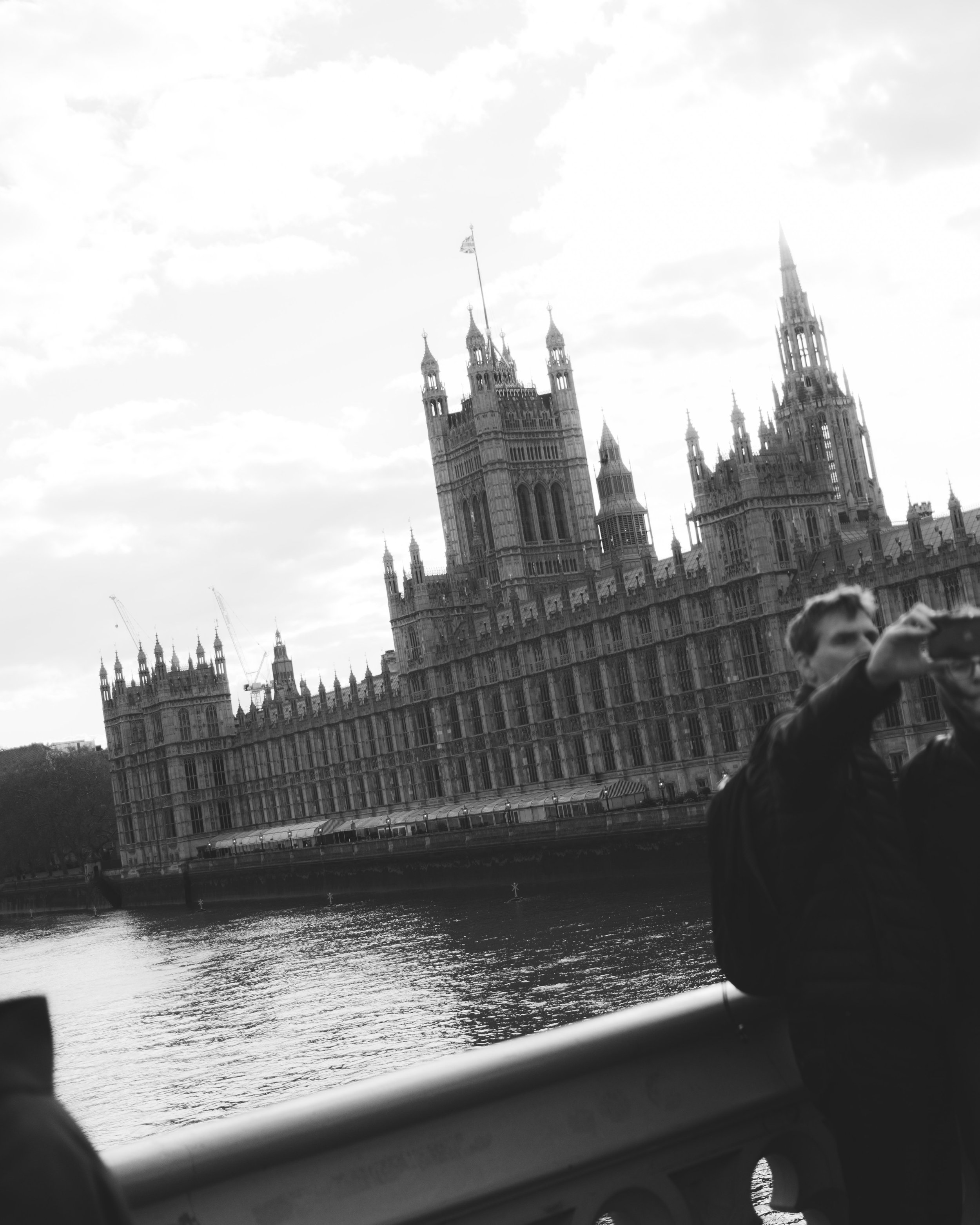 Black and white photo of the Houses of Parliament and Big Ben in London, viewed across the river, with clouds in the sky. Part of a person taking a selfie or photo with the buildings in the background.