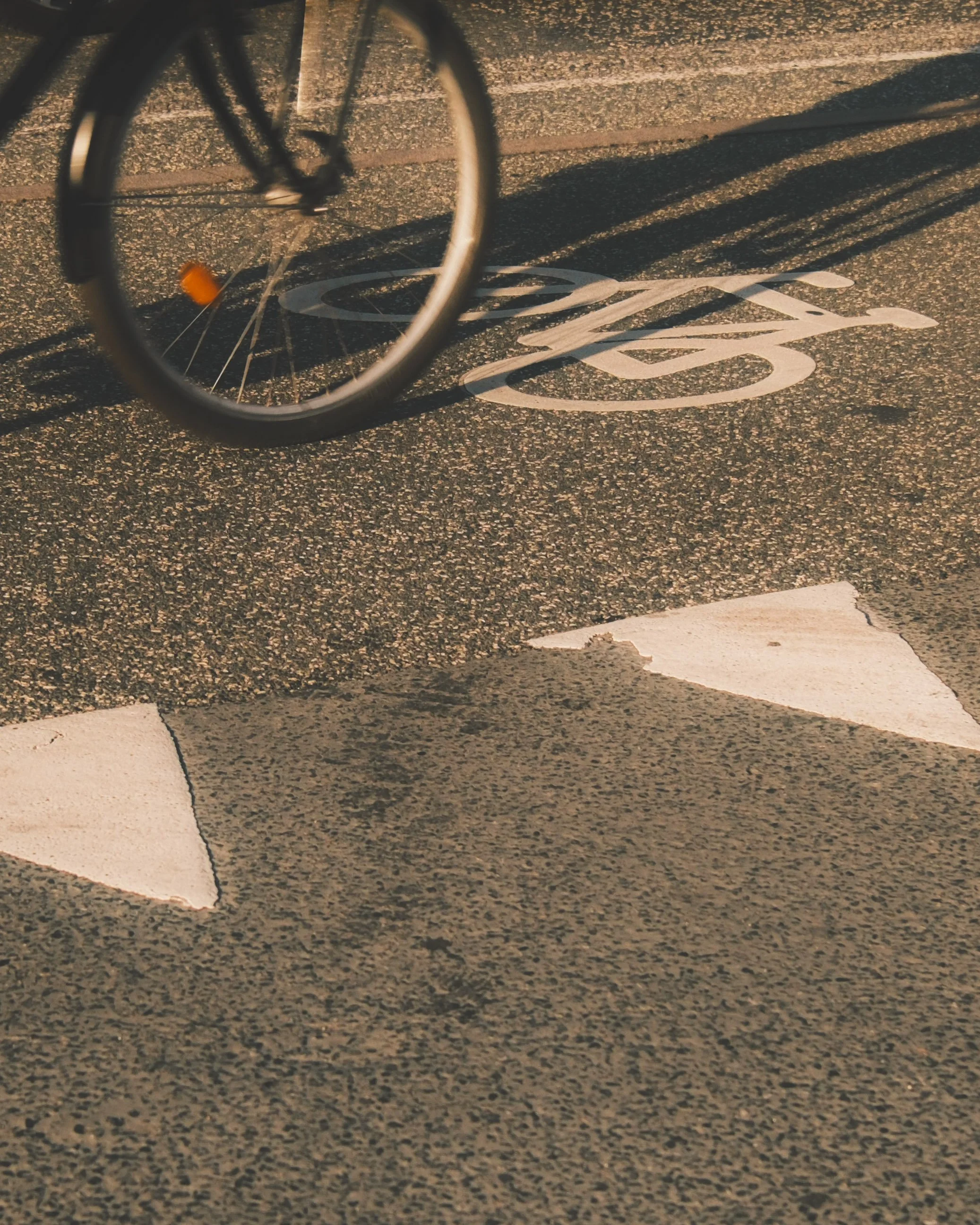 A bicycle wheel casting a shadow onto a textured asphalt bike lane with a white bike lane arrow.