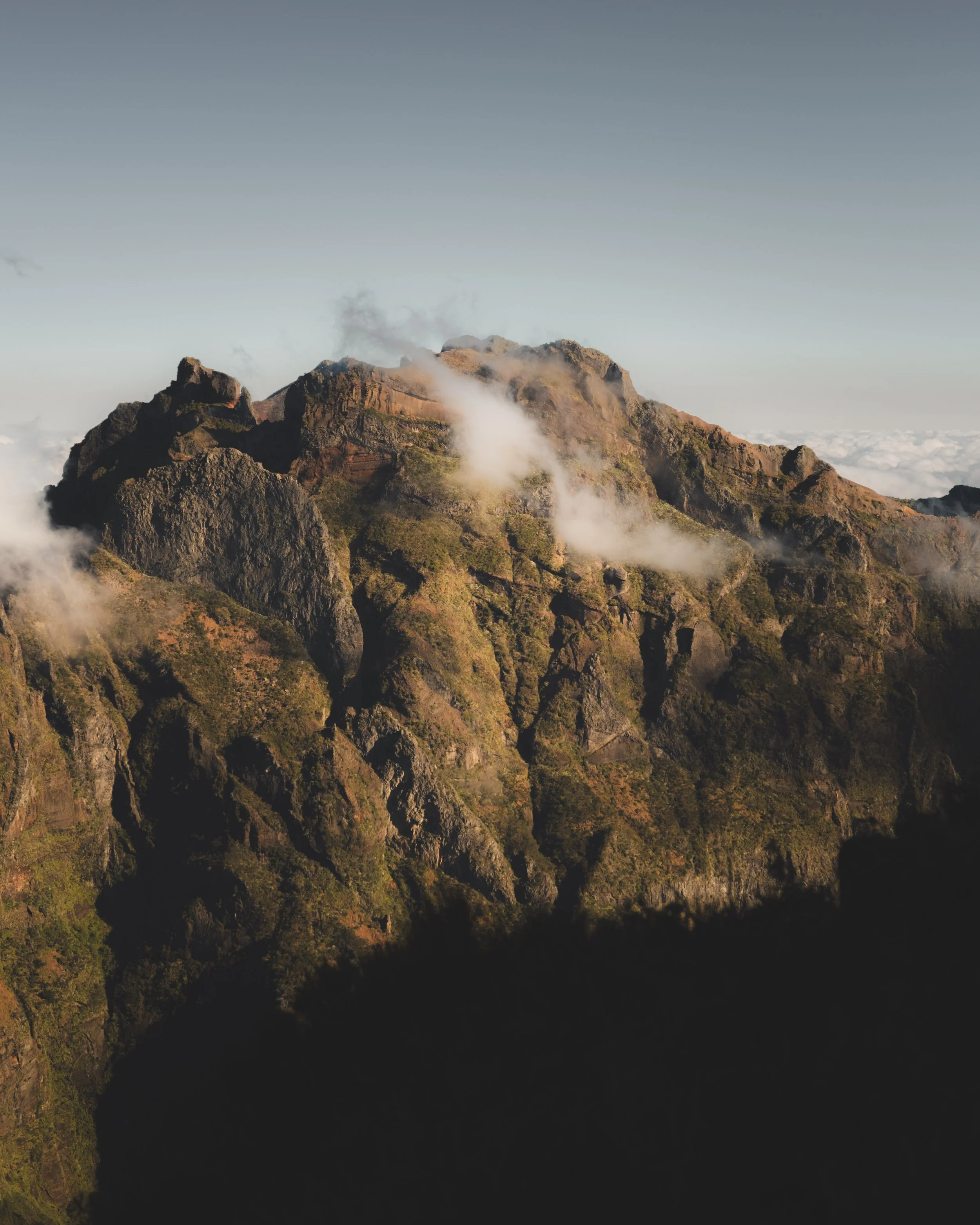 Sunlit rugged mountain range with mist and clouds around the peaks, viewed during daylight hours.