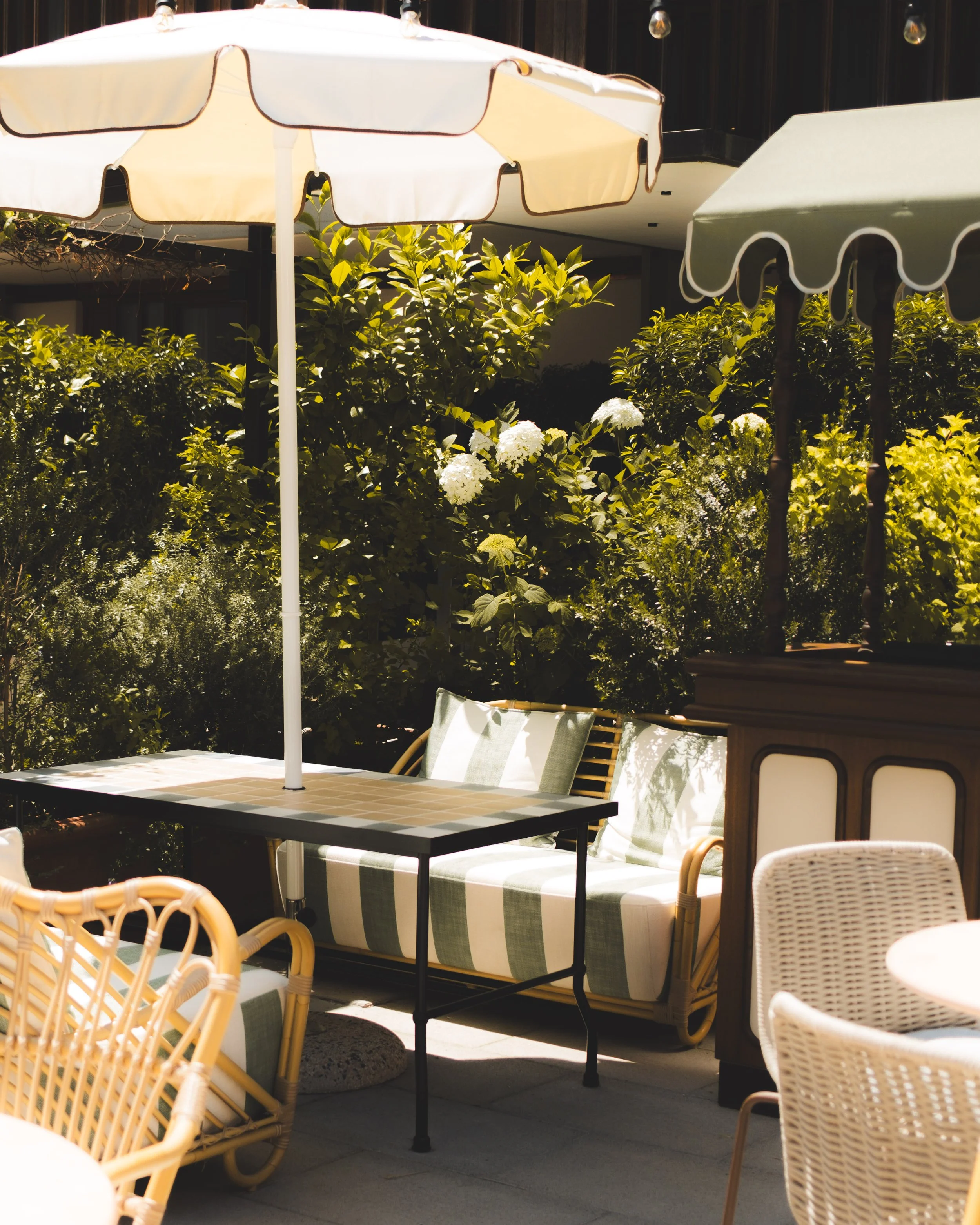 An outdoor patio area with beige and green striped cushioned lounge chairs and tables under large cream-colored umbrellas, surrounded by greenery and white hydrangea flowers.