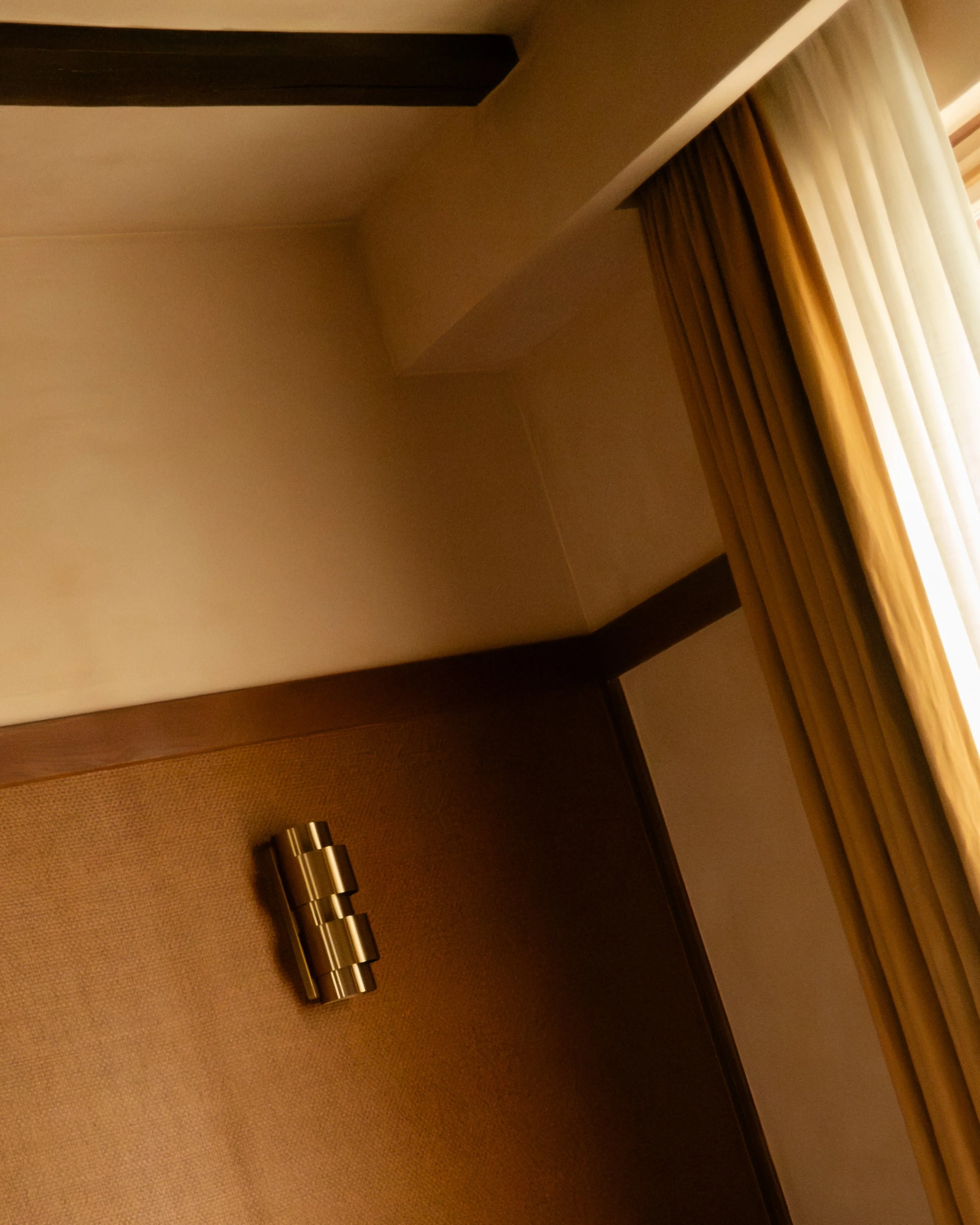 Interior scene showing a corner of a room with beige walls, wooden paneling, and window curtains. There is a metallic hinge or hinge-like object on the beige carpeted floor. Lionel Chu - Hotel & Travel Photography