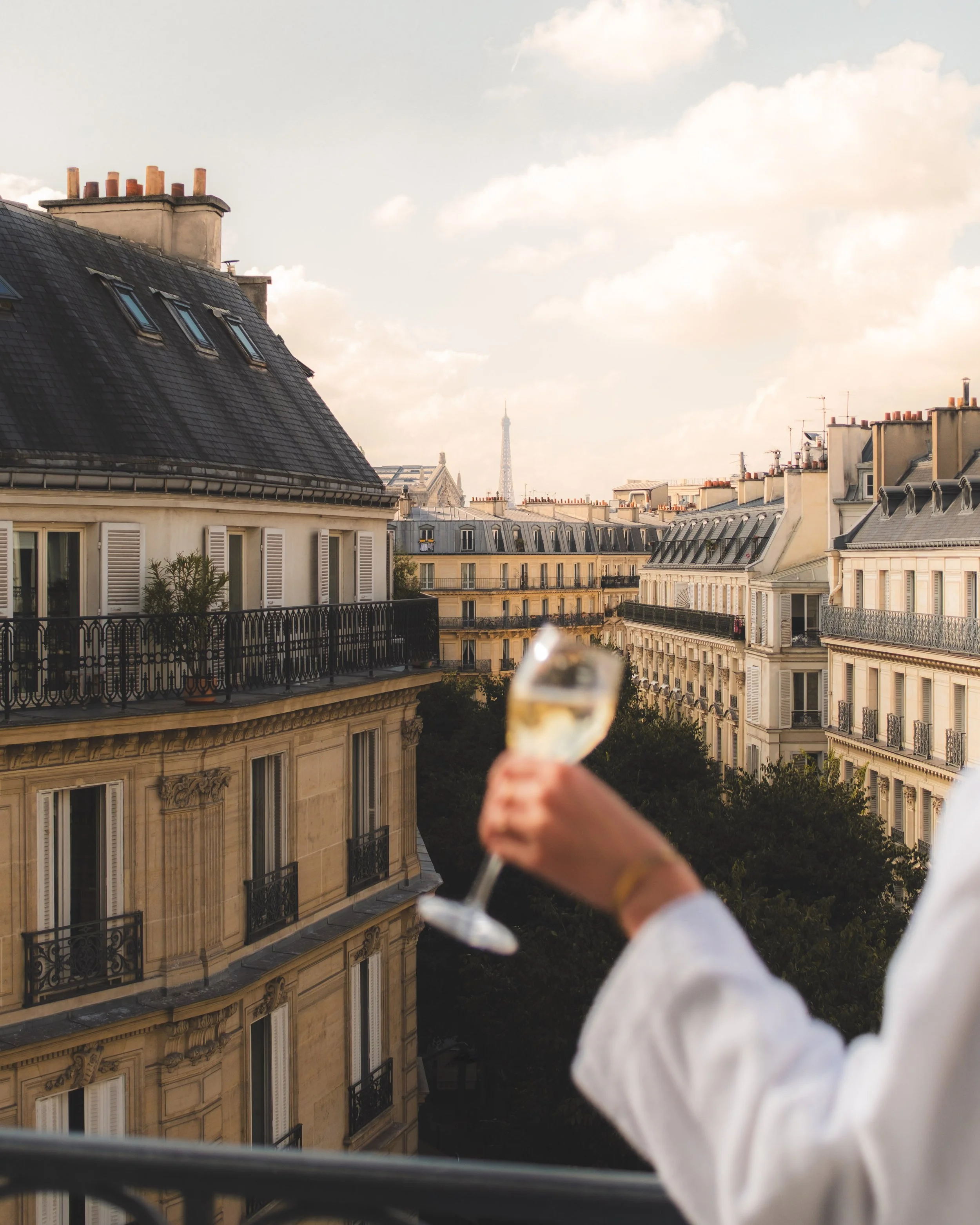 A view of Paris rooftops with the Eiffel Tower in the distance, a person is holding a glass of white wine in the foreground.