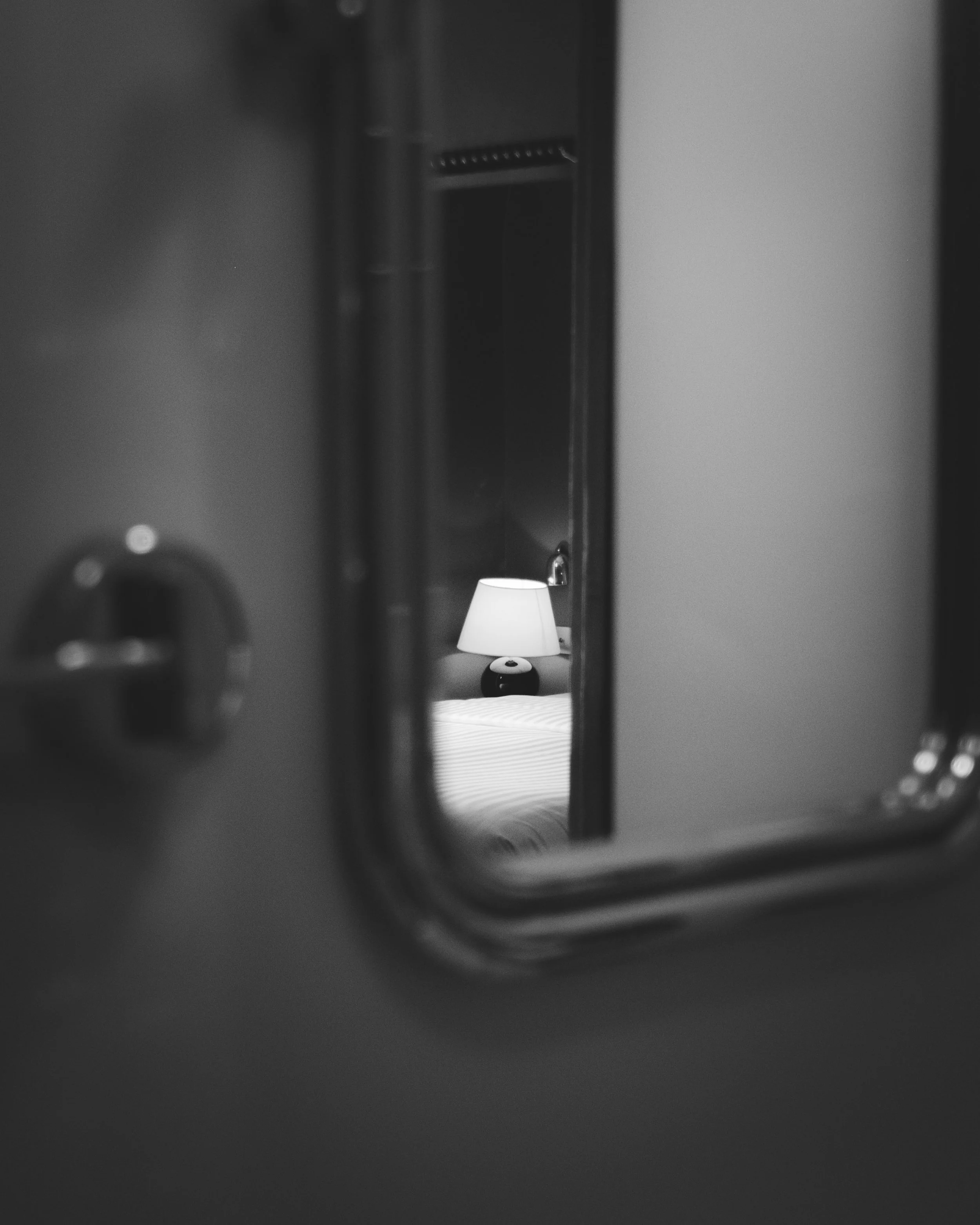 View through a small opening showing a bedside lamp with a white shade on a nightstand, reflected in a mirror in a bedroom