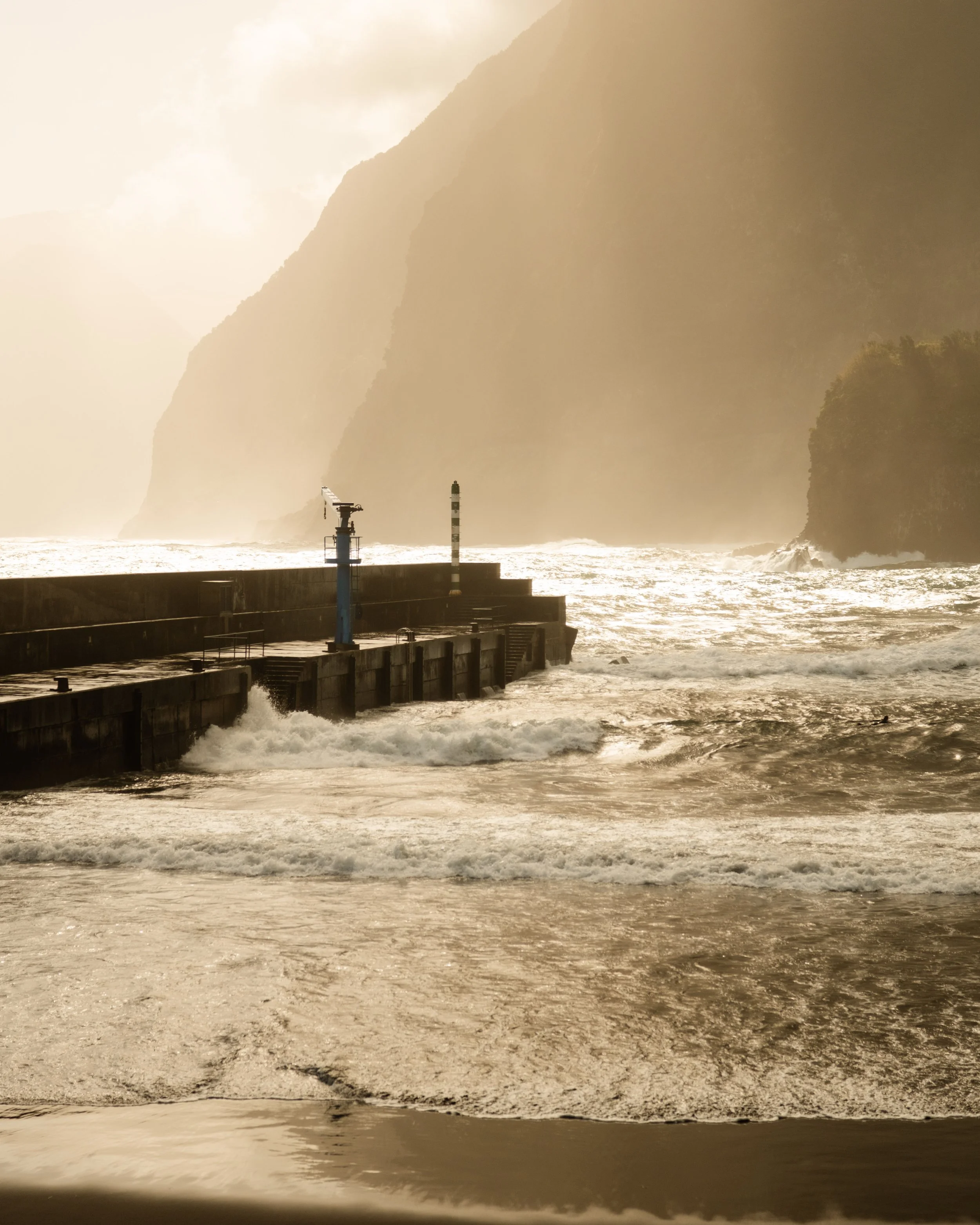 A coastal scene with a breakwater pier, a warning light, large waves, and a foggy atmosphere with mountains in the background.