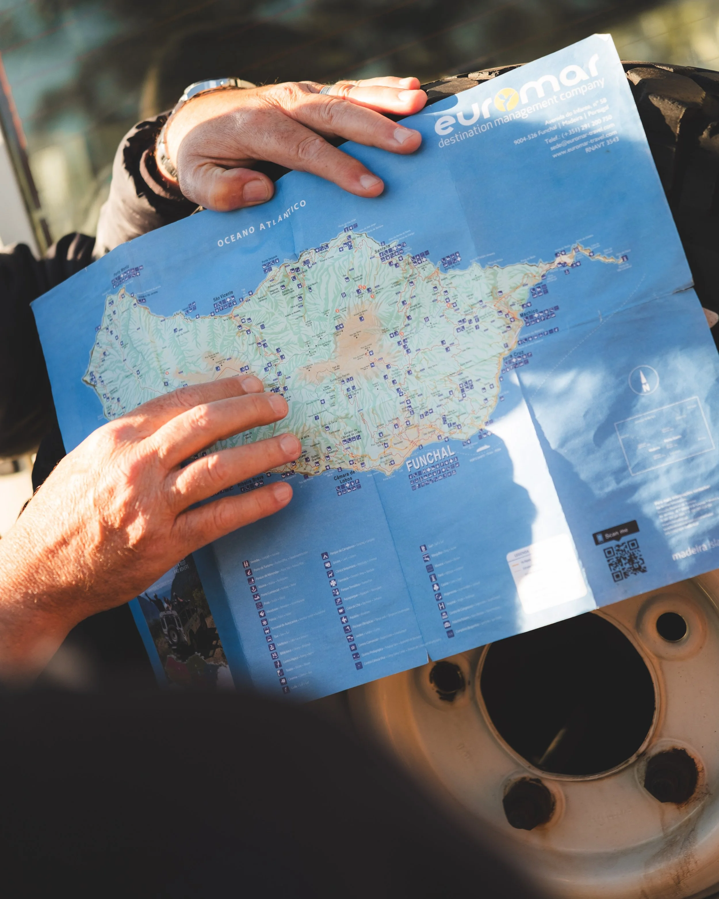 A person is holding a map of the Funchal area in Portugal, with a hand on the coast near the Atlantic Ocean, standing over a circular, rusted car wheel rim.