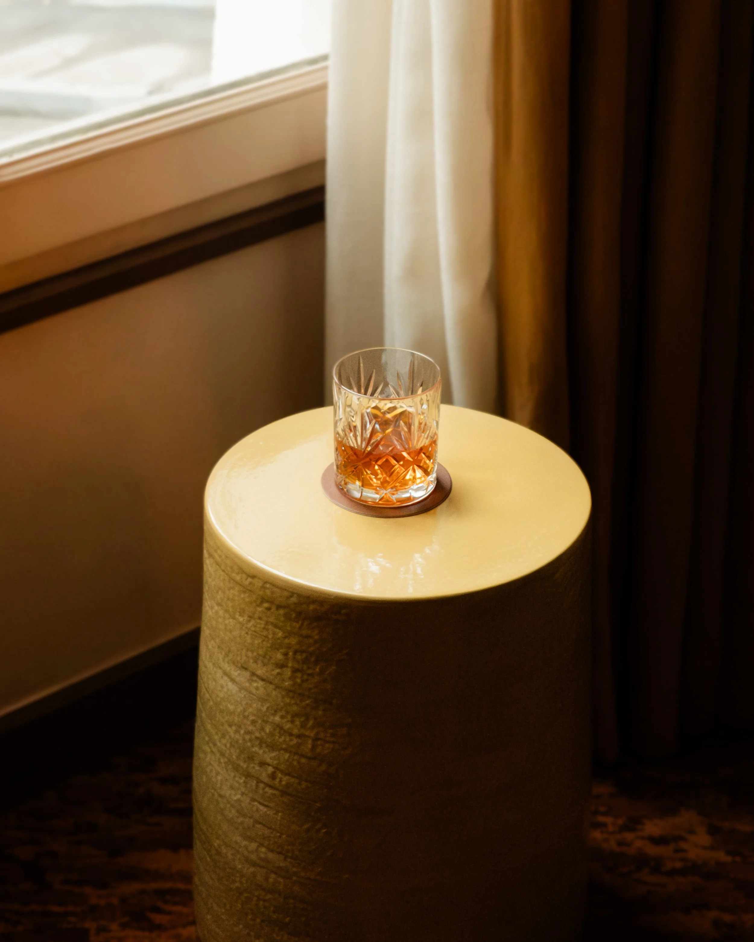 A glass of whiskey on a round beige table next to a window with curtains. Lionel Chu - Hotel & Travel Photography