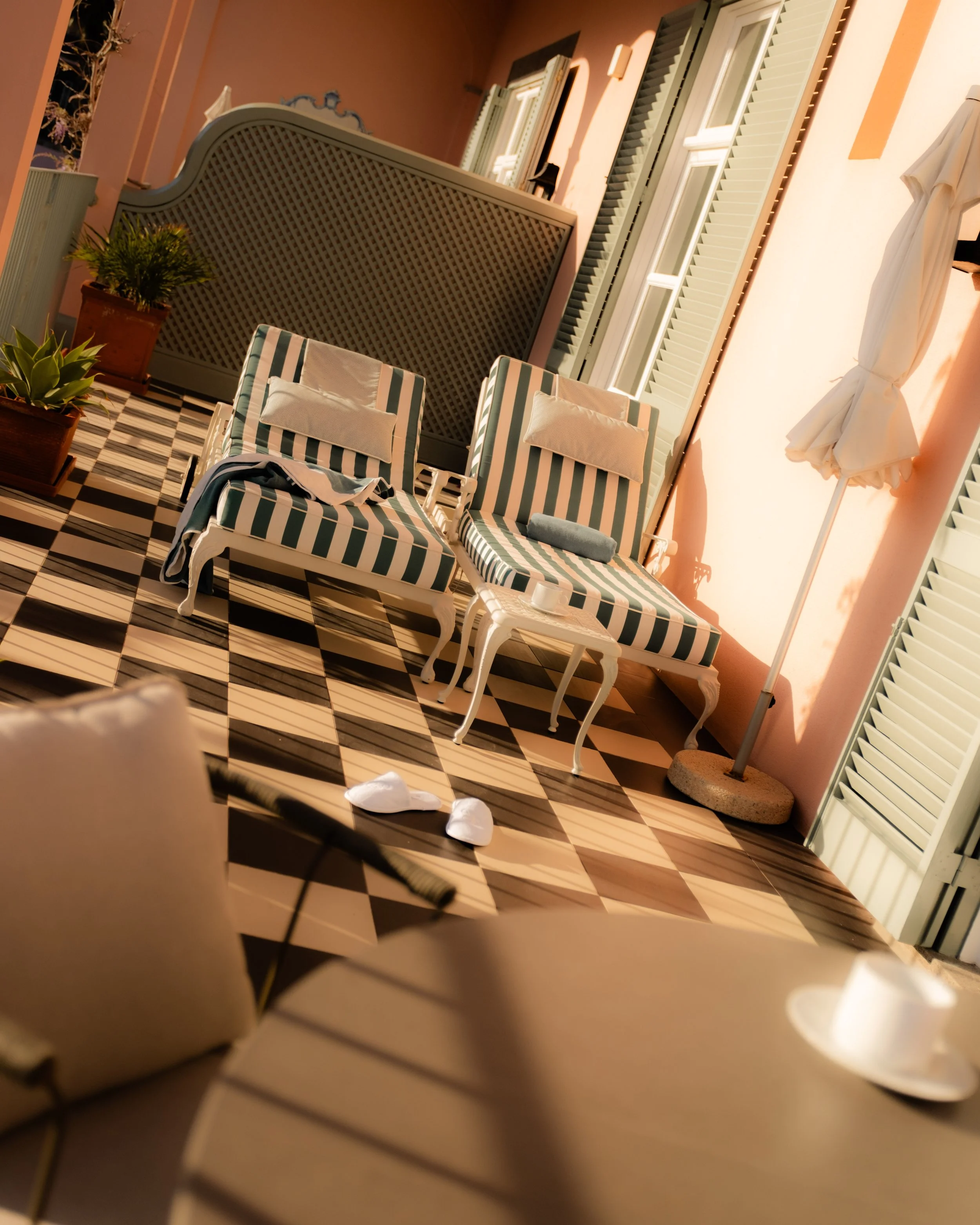 A sunlit outdoor patio with two white lounge chairs with green and white striped cushions, a small puff of clothing on one chair, various potted plants, a pink wall, and closed green shutters on windows.